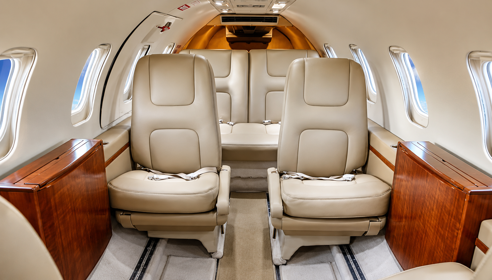 Interior of a private jet with beige leather seats, windows, and wooden storage cabinets.