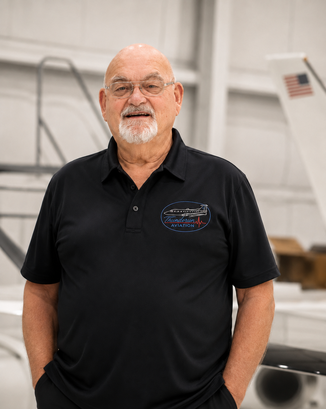 An older man with glasses, a white beard, and a bald head, wearing a black polo shirt with a logo that says 'Thundergun Aviation', standing inside an aircraft hangar.