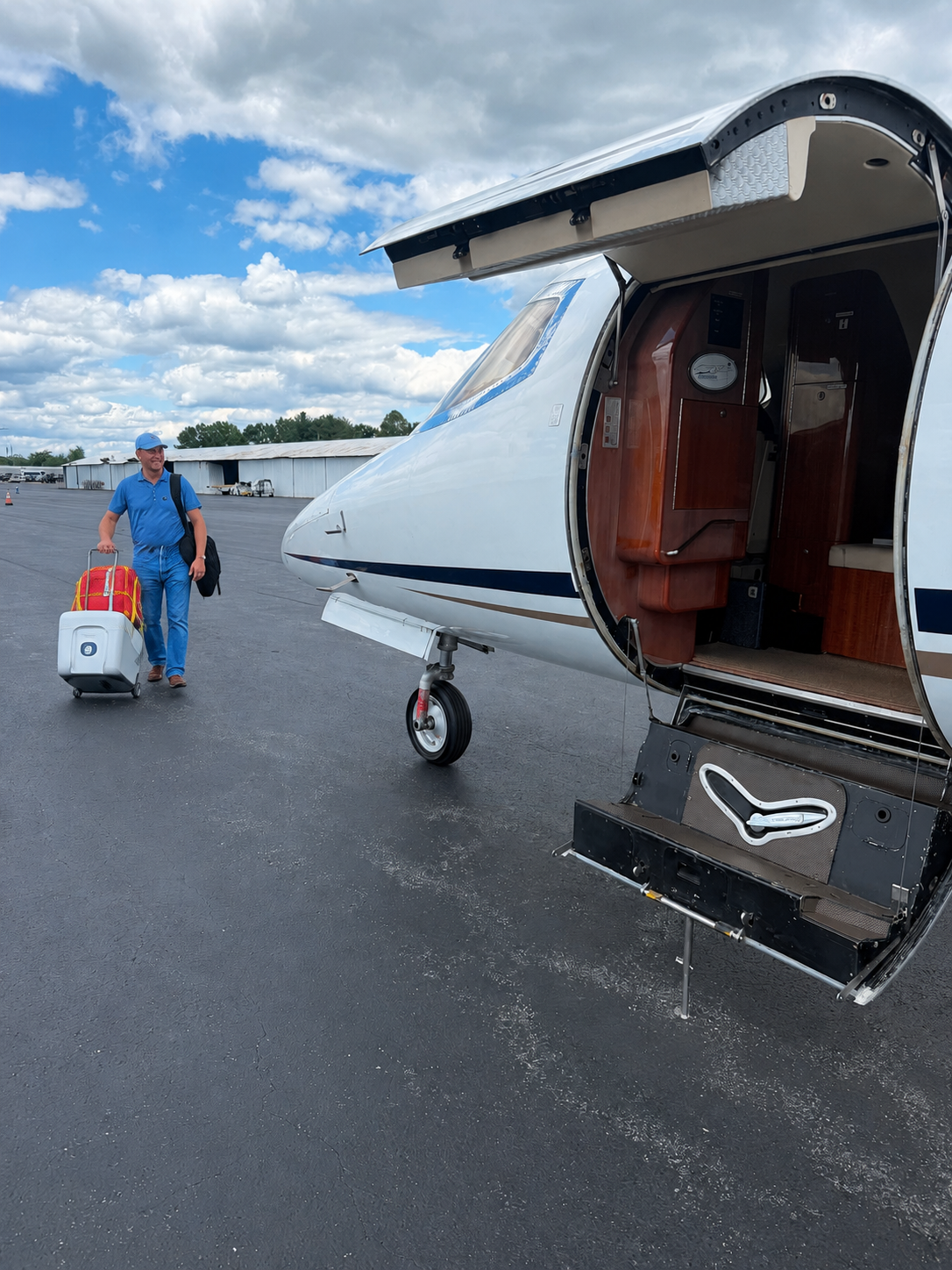 Man walking on tarmac pulling a rolling suitcase away from a small private jet with its door open, showing wood interior.