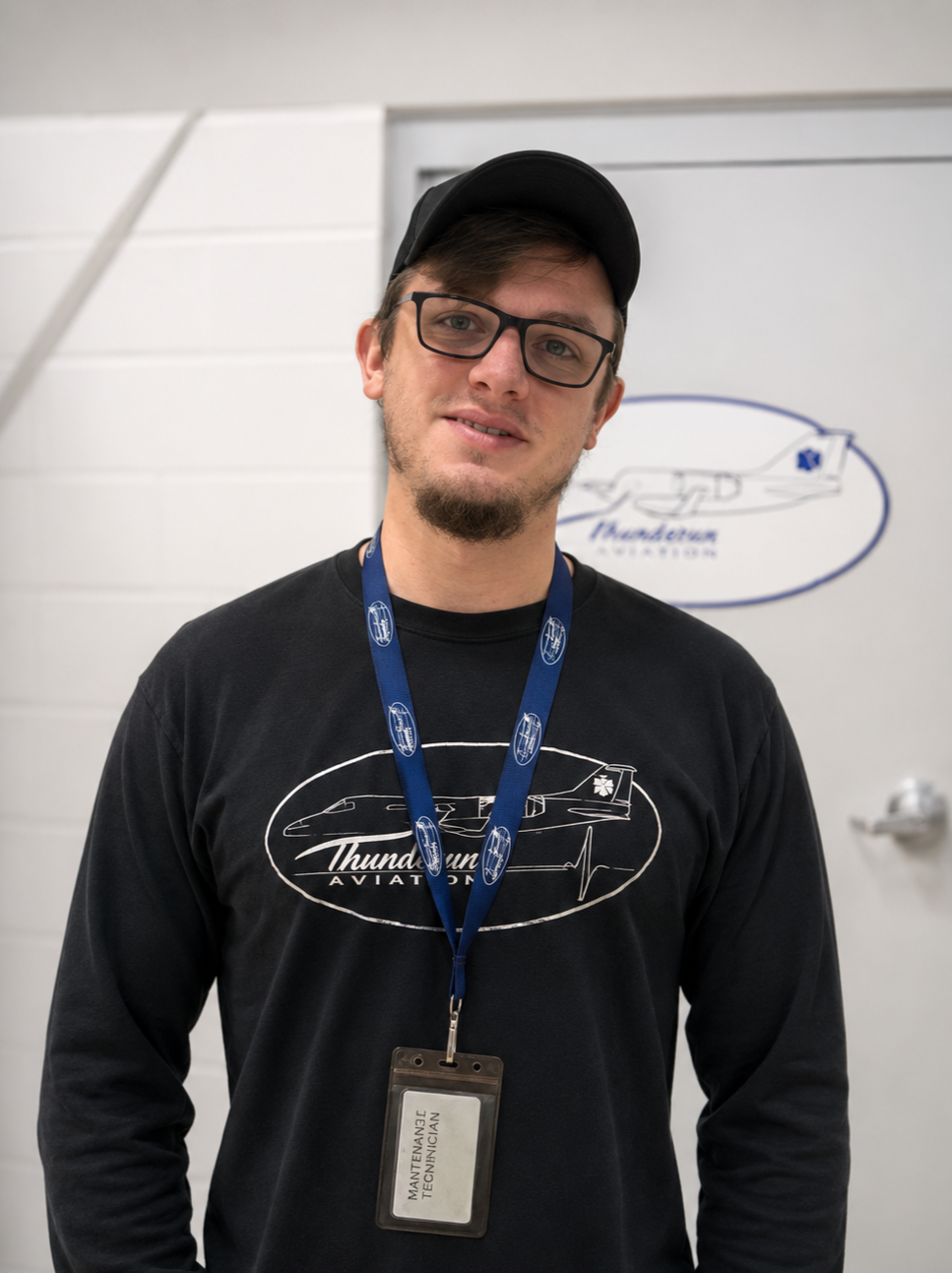 A young man with glasses, a baseball cap, and a beard, wearing a Thundrums aviation sweatshirt and a maintenance technician badge, standing indoors in front of a whiteboard with a drawing of an aircraft.