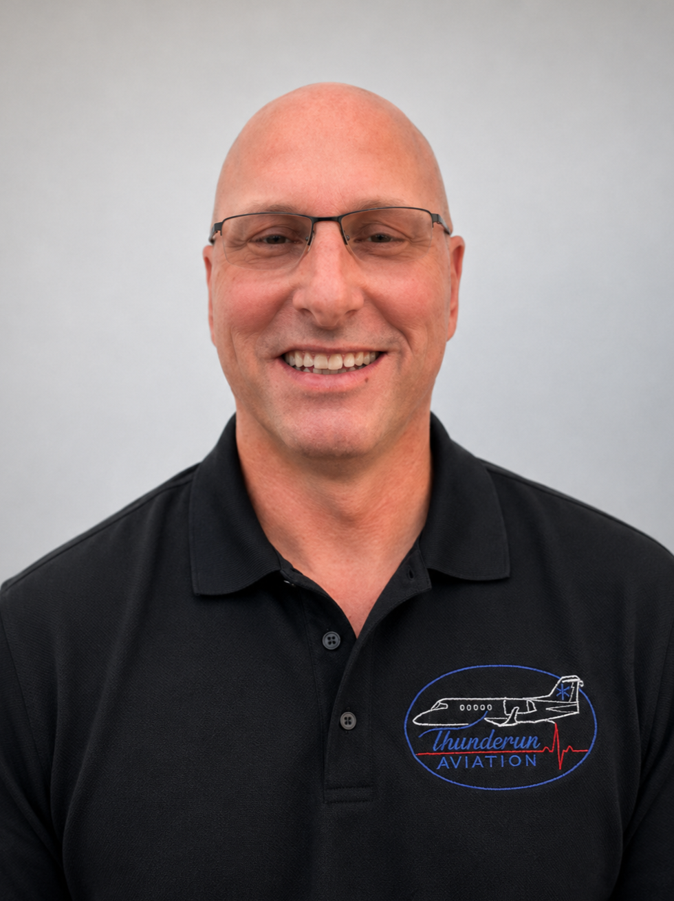 A smiling man with glasses wearing a black polo shirt with a ThunderRun Aviation logo, standing against a plain light gray background.