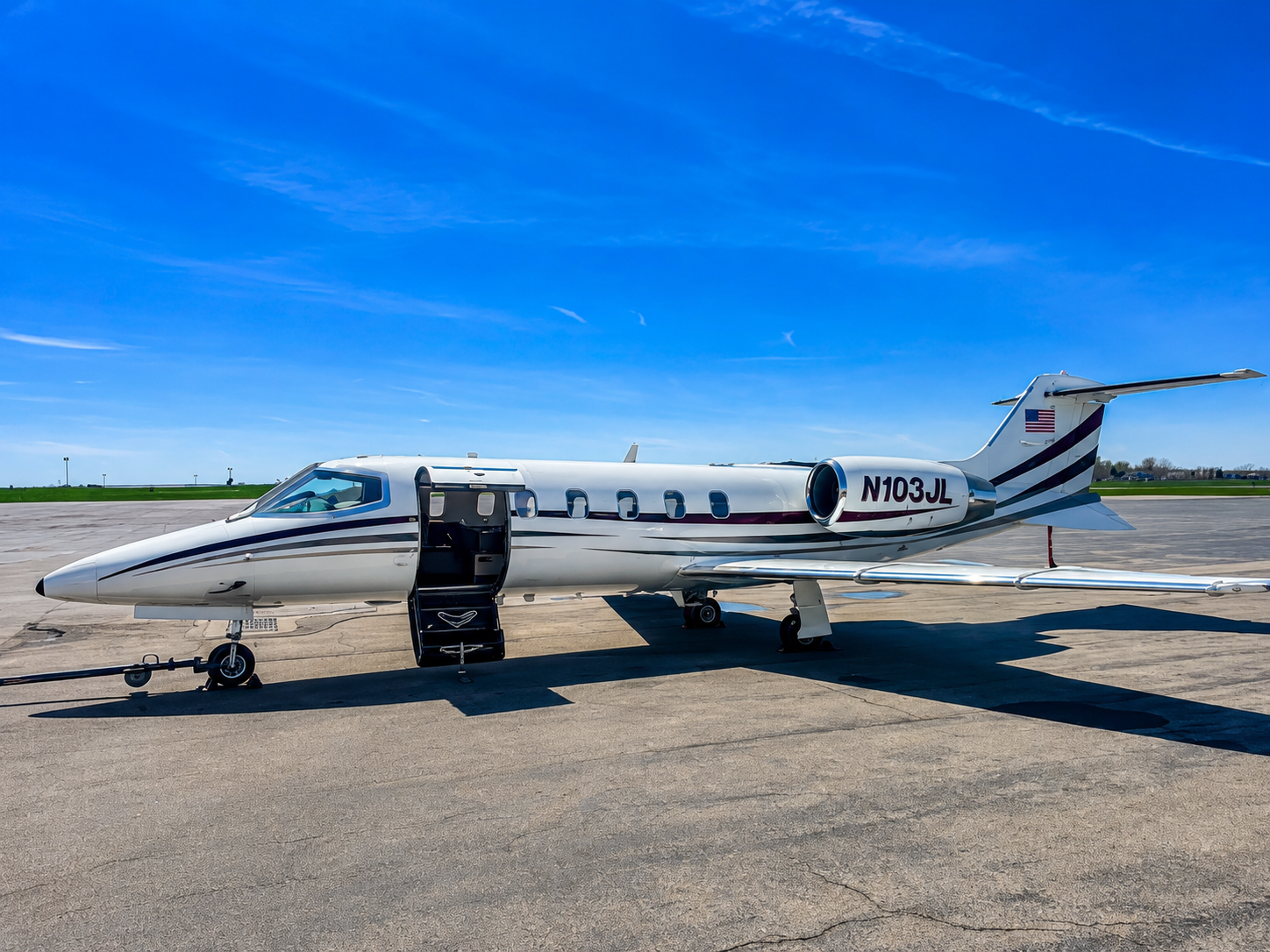 A private jet on the tarmac under a clear blue sky, with its door open and steps extended for boarding.