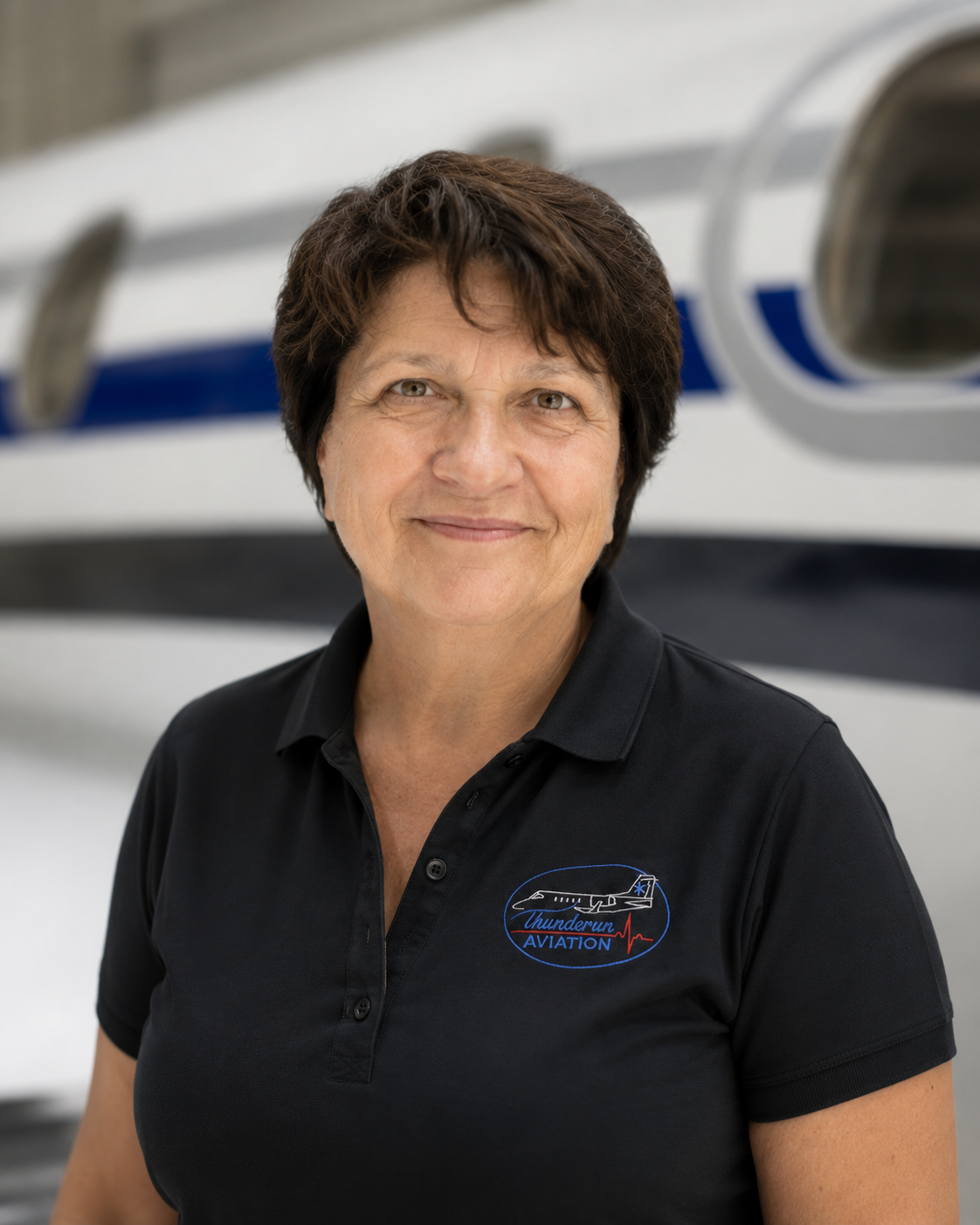 A woman with short dark hair smiling in front of a small private jet, wearing a black polo shirt with a logo that reads 'Thundermun Aviation' and an airplane graphic.