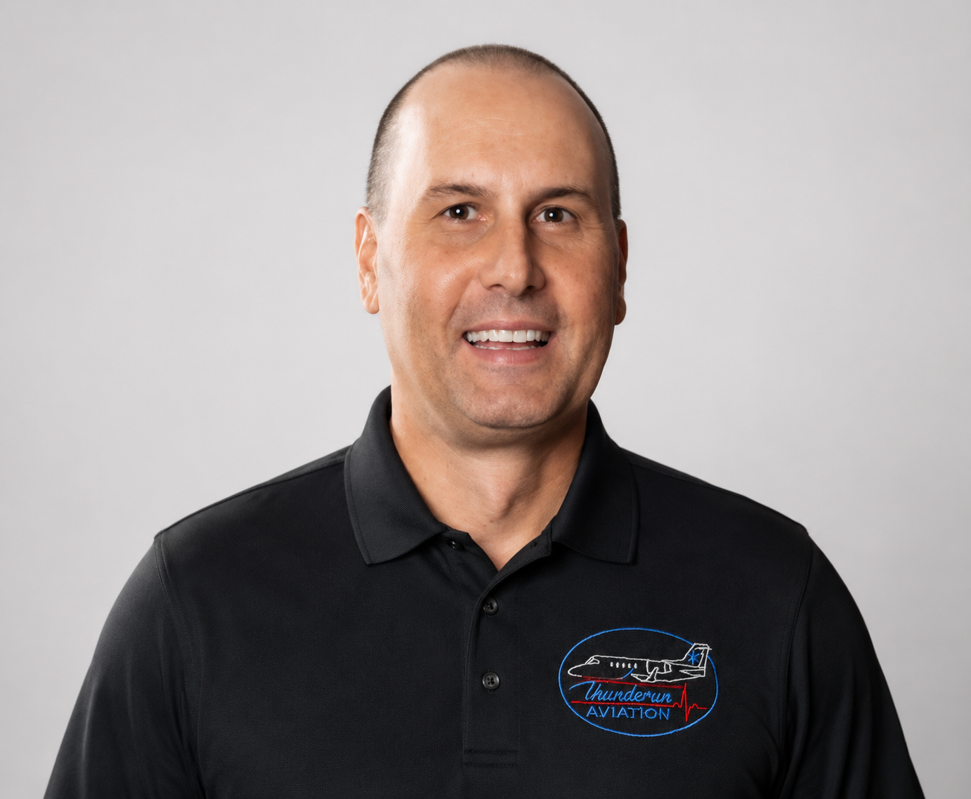 A man with a shaved head smiling, wearing a black polo shirt with a Thundervan Aviation logo on the chest, standing against a plain light gray background.