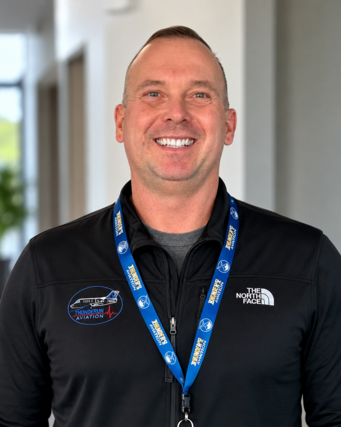 Smiling man in black jacket with 'Thunder Run Aviation' logo and North Face badge, wearing a blue lanyard, indoors with a blurred background.