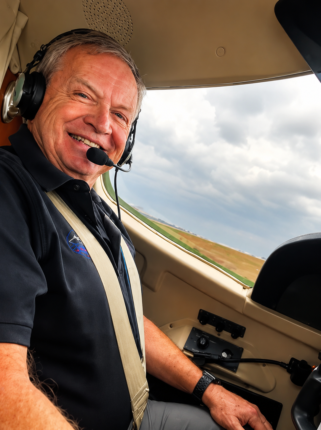 A smiling man sitting in the cockpit of an aircraft with a headset on, in front of a cloudy sky and open landscape.