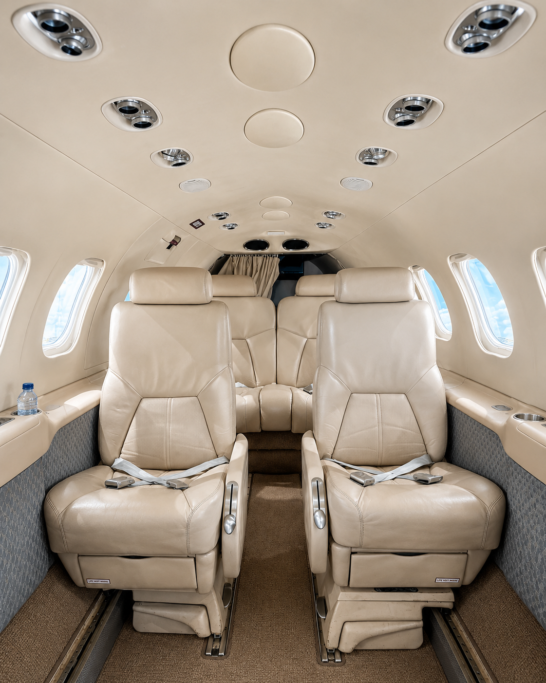 Interior of a private jet with beige leather seats and multiple windows, ceiling air vents, and small personal space compartments.