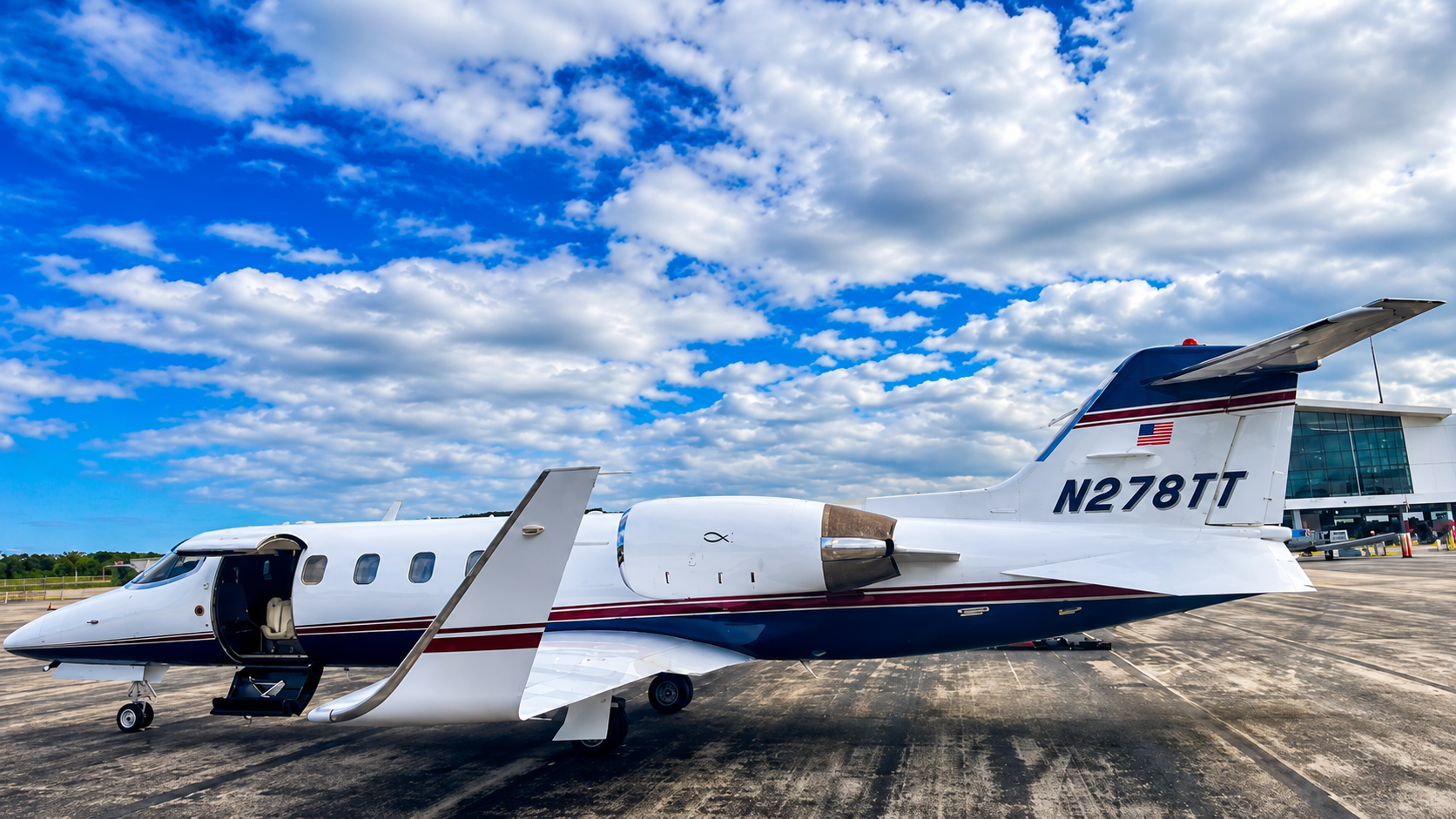 Private jet parked on tarmac near airport terminal, under partly cloudy blue sky.