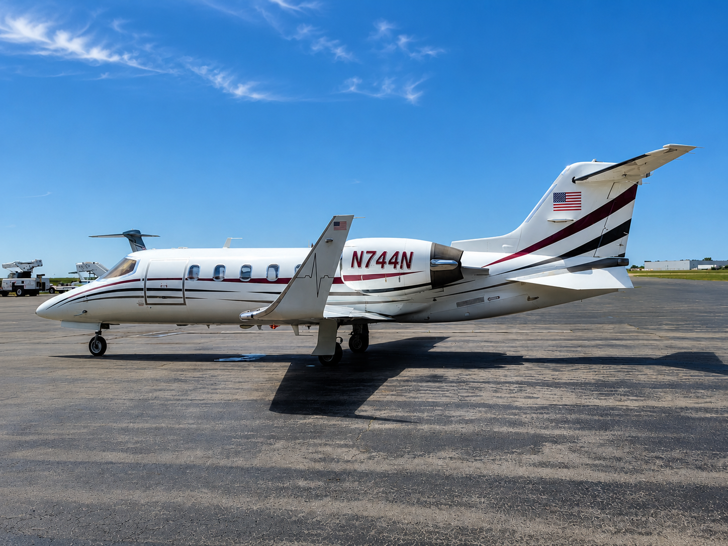 White private jet with red and black stripes parked on tarmac under blue sky, with ground vehicles in background.