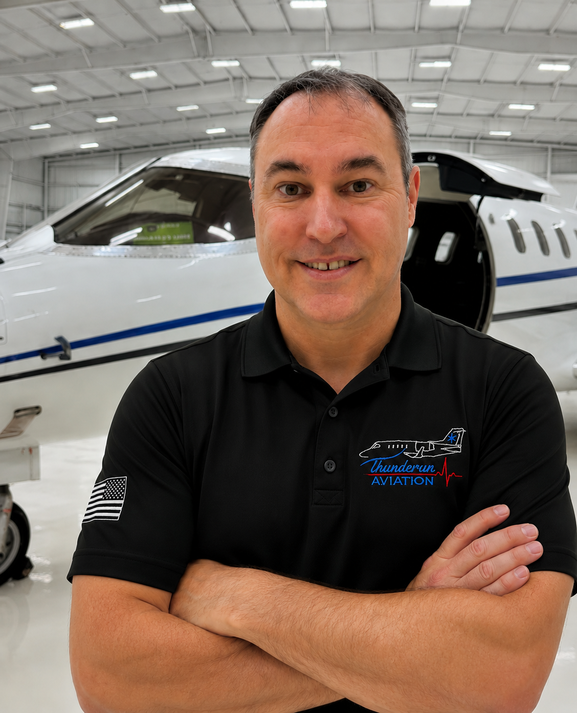 A man with crossed arms smiling in front of a private jet inside an aviation hangar, wearing a black polo shirt with Thundrun Aviation logo.