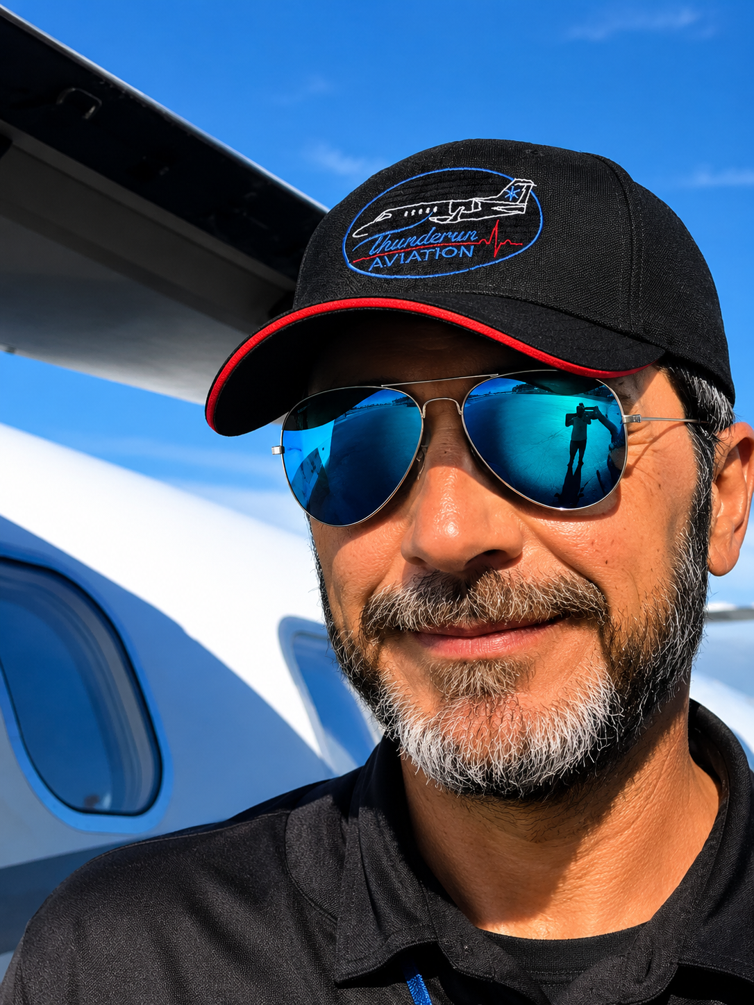 A man wearing sunglasses and a black cap with a logo, standing in front of a private jet, taking a selfie.