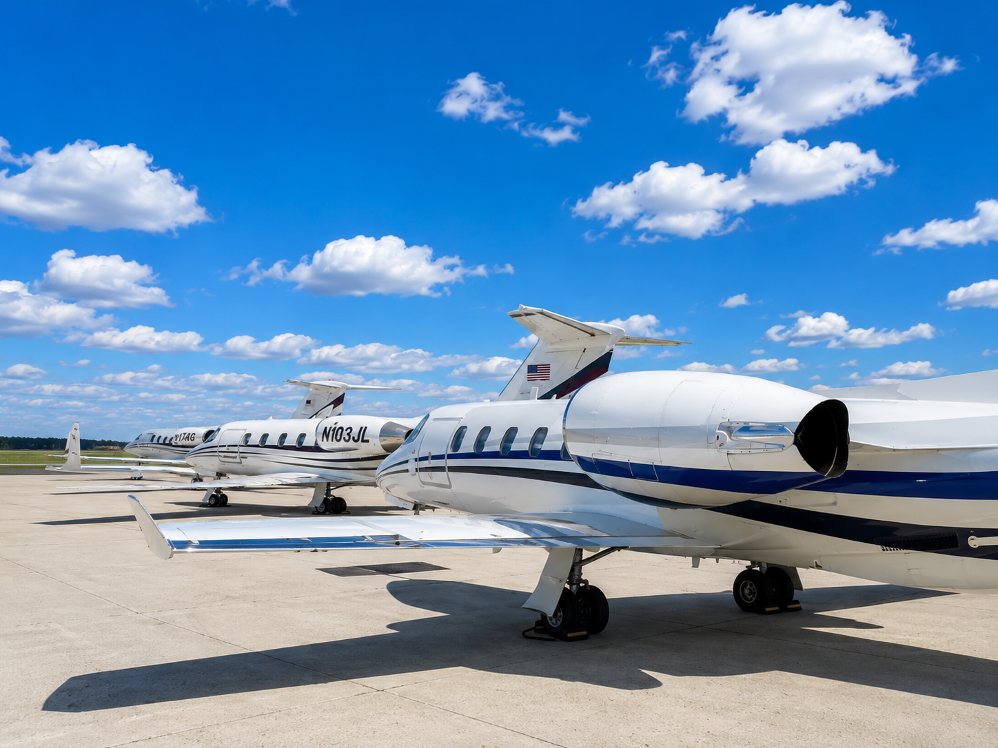 Three private jets parked on an airport tarmac under a blue sky with scattered clouds.