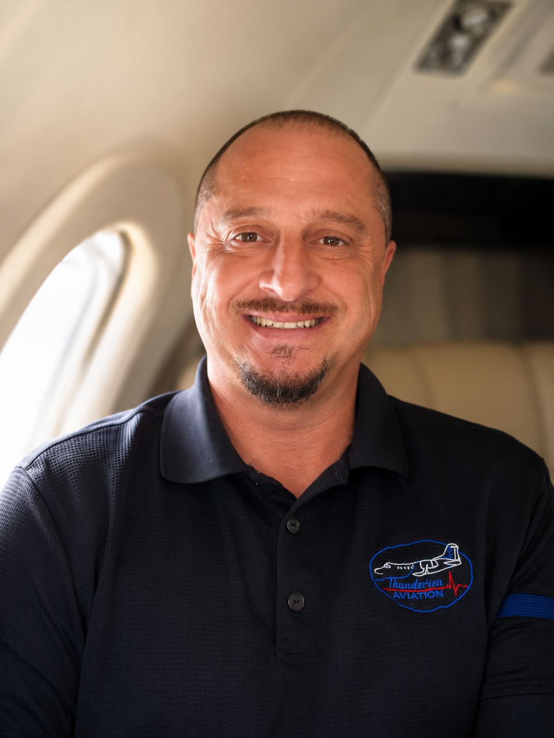 A smiling man with short hair, a goatee, and wearing a black polo shirt with a logo that reads 'ThunderView Aviation' sitting inside an airplane.