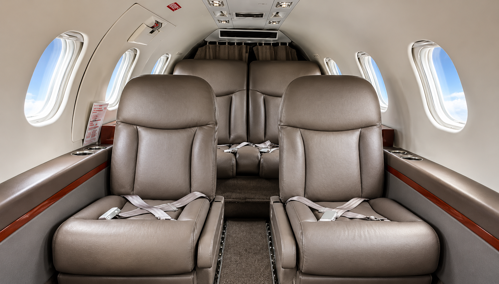 Empty airplane seats with beige leather upholstery and seat belts inside a cabin, with windows showing clouds and sky.
