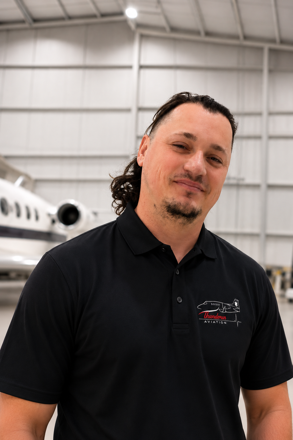 A man with long dark hair, a goatee, and a slight smile standing inside an aircraft hangar, wearing a black polo shirt with an aviation logo.