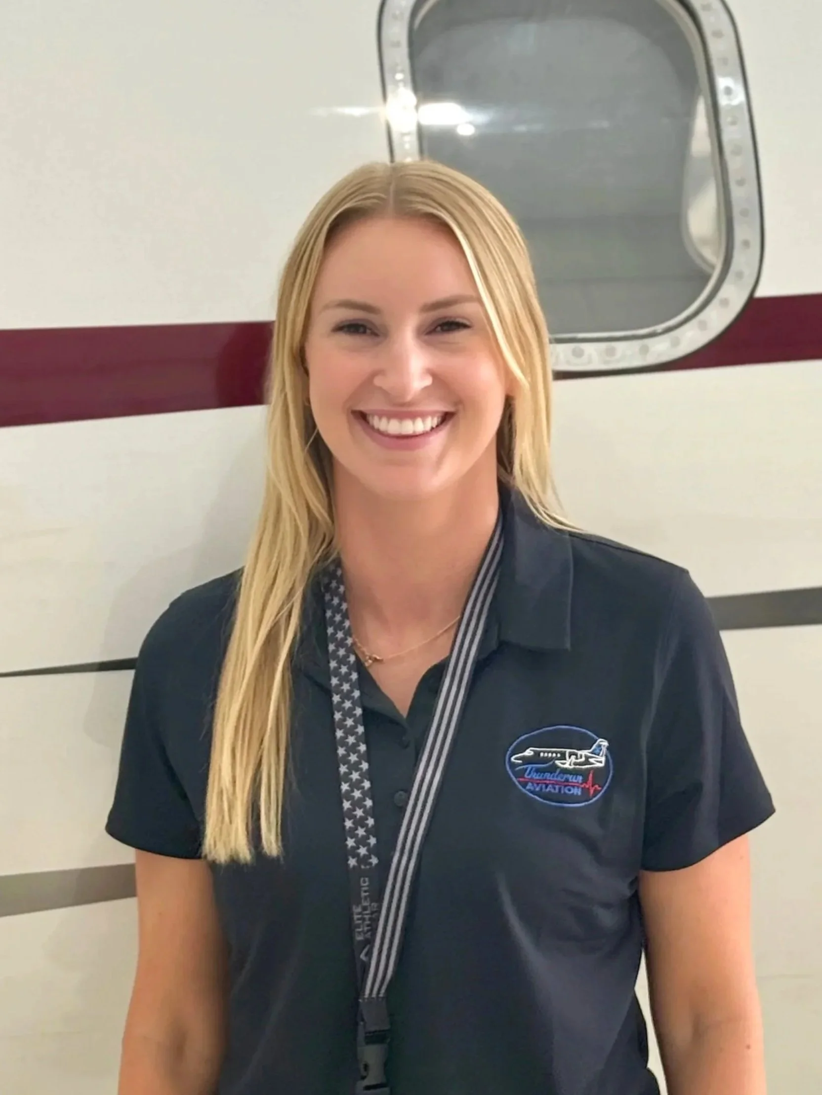 A woman with long blonde hair wearing a black polo shirt with the Thunderbirds aviation logo, standing in front of an aircraft.