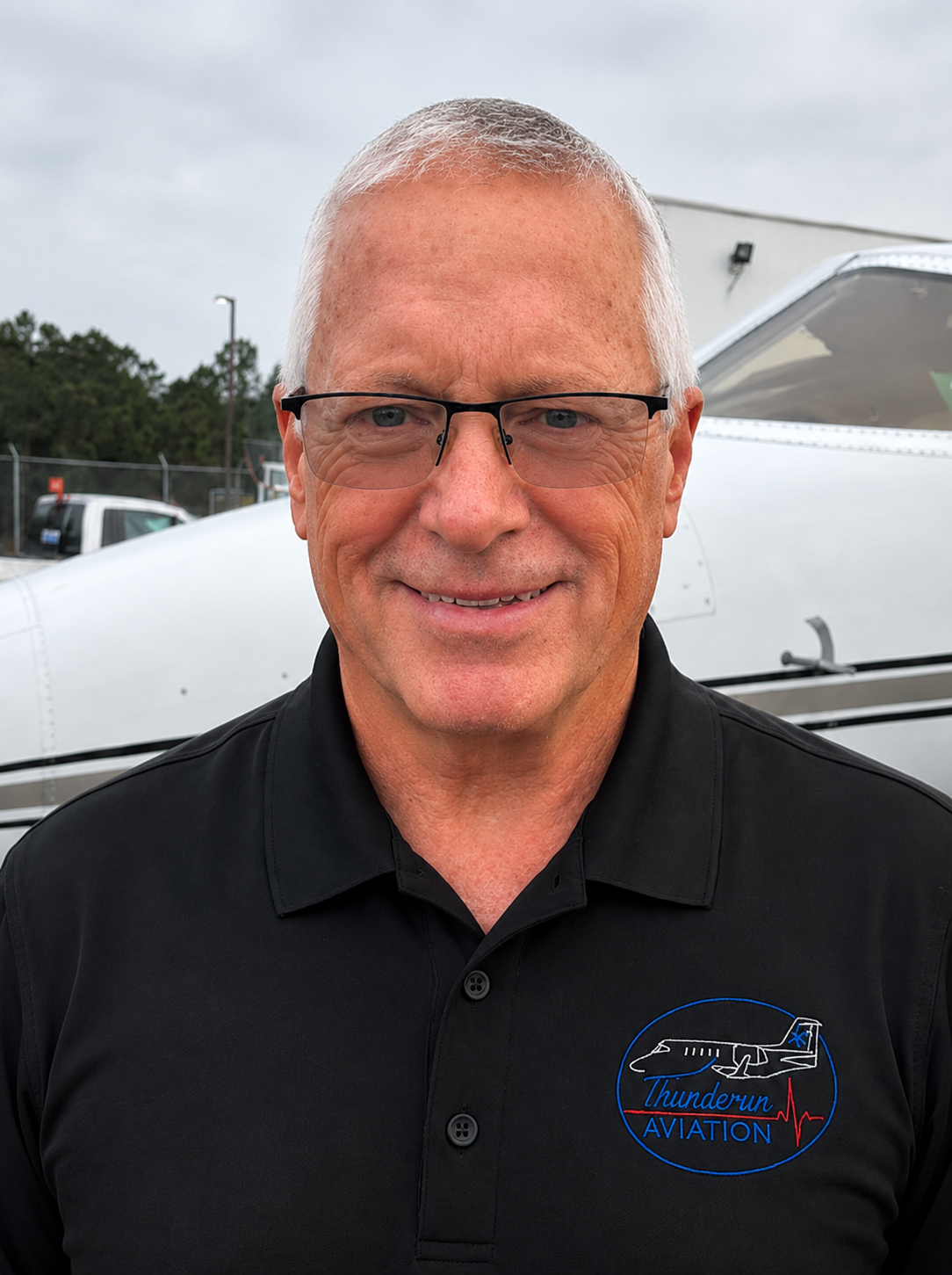 A smiling man with short gray hair, glasses, wearing a black polo shirt with a Thunderun Aviation logo, standing in front of a small aircraft on a cloudy day.