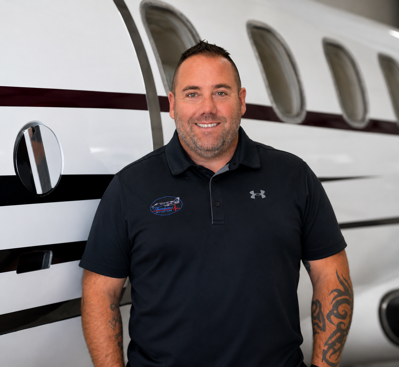 A smiling man with a beard and short hair stands in front of an airplane, wearing a black Under Armour shirt with an FAA patch.