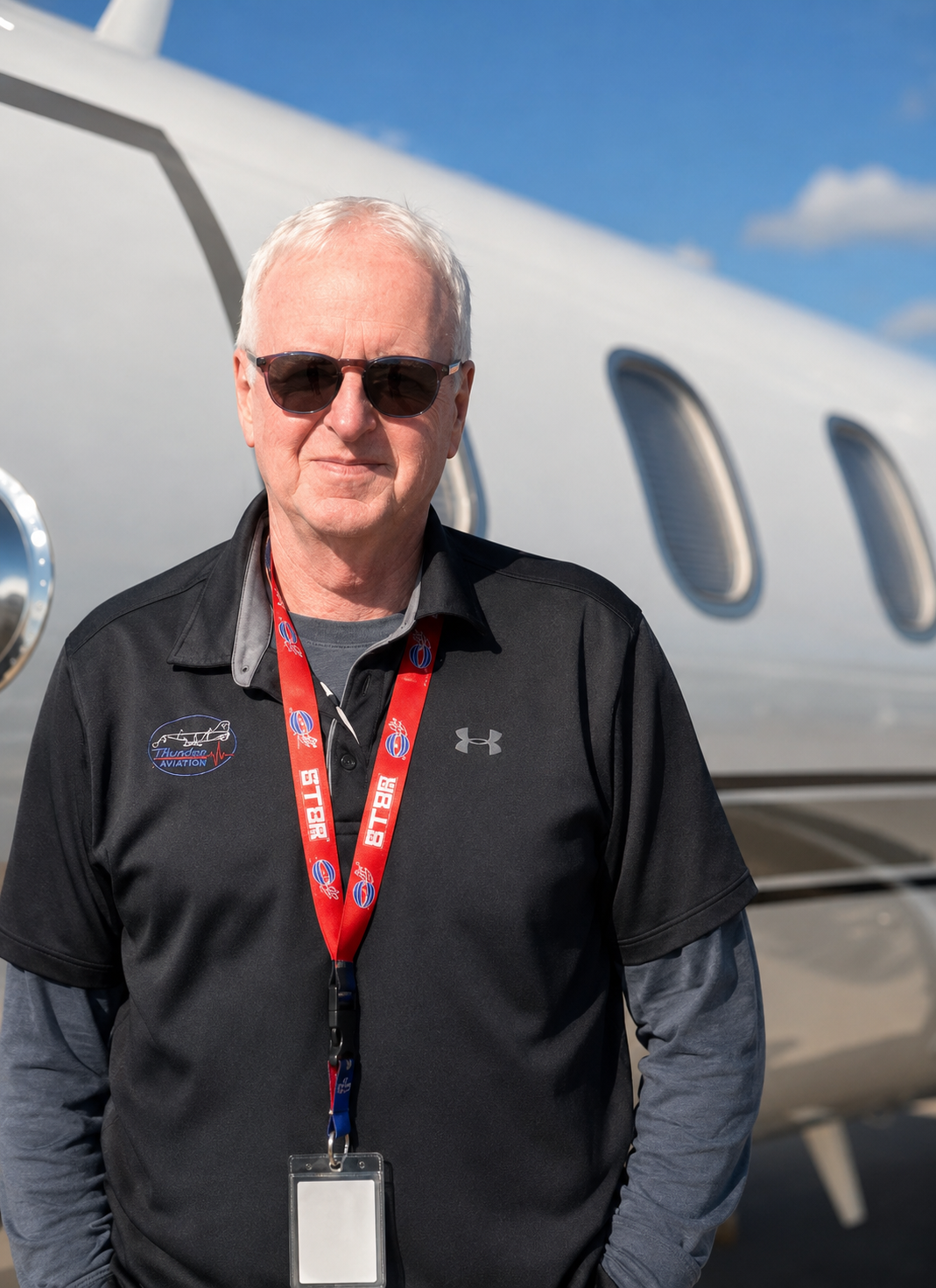 An older man wearing sunglasses, a black Under Armour polo with an aviation logo, a red lanyard, and a gray long-sleeve shirt underneath, standing in front of a private jet on a clear day.