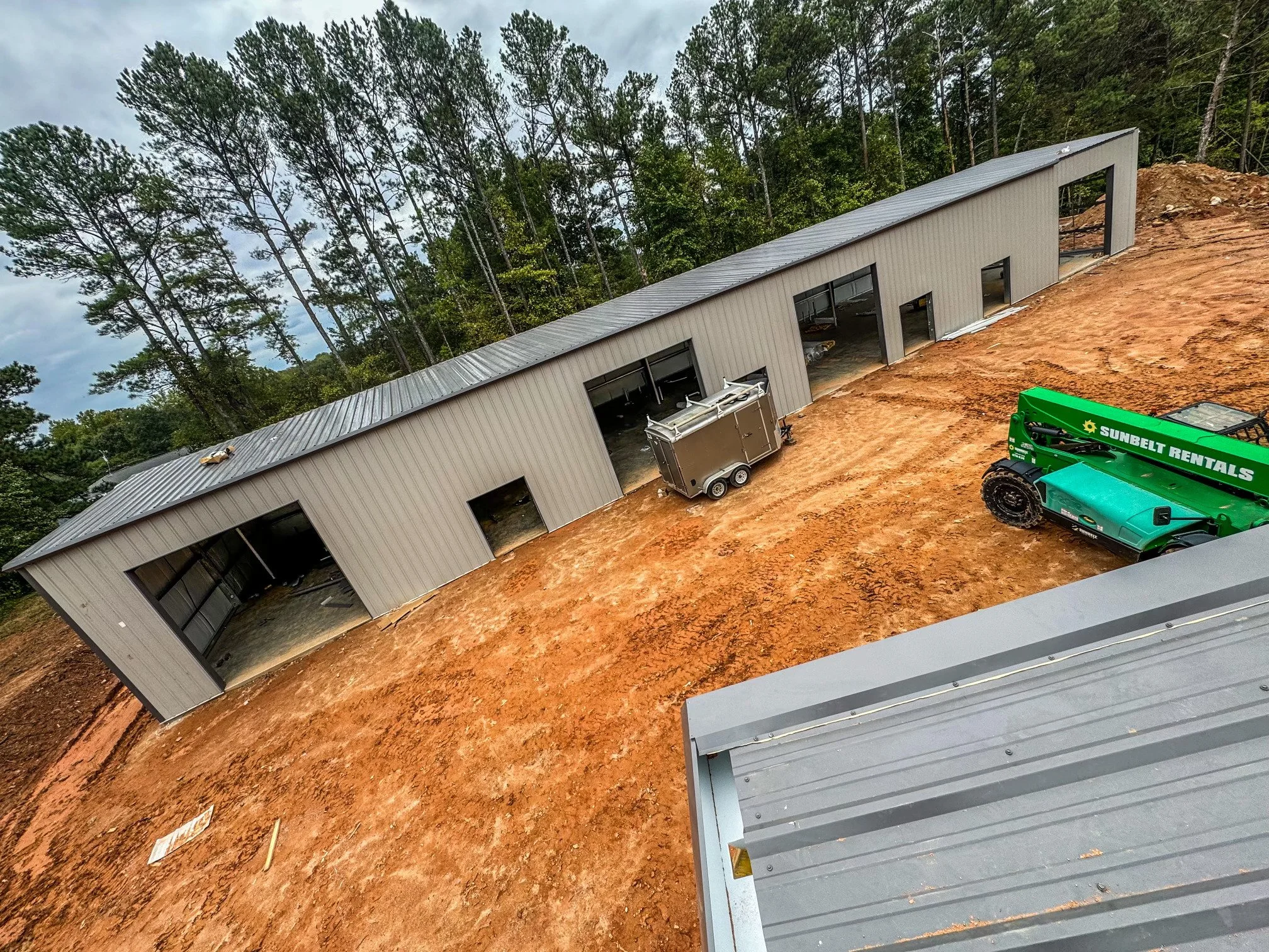 New metal building under construction on dirt ground with construction equipment, including a green Sunbelt Rentals vehicle and a silver trailer, surrounded by a forested area.