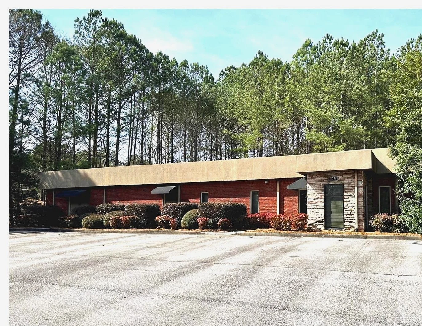 A single-story commercial building with a flat roof and brick facade, surrounded by a parking lot and landscaped bushes, with trees in the background.