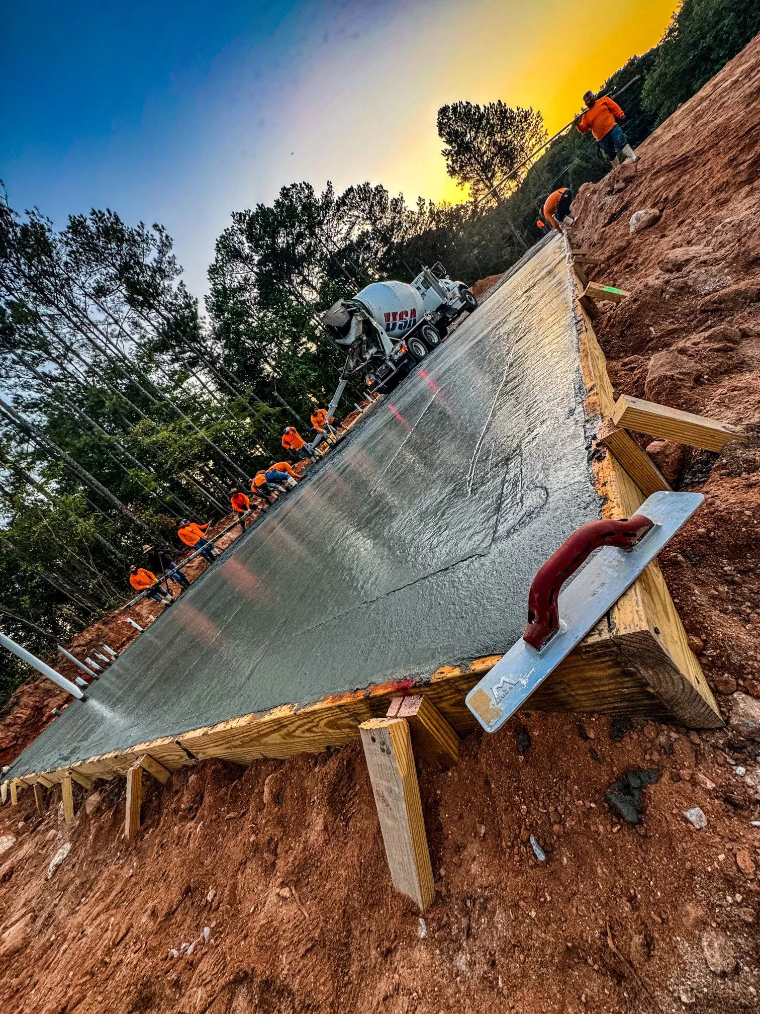 Construction workers building a concrete slab on a wooden form at sunset, with a cement mixer truck in a forested area.