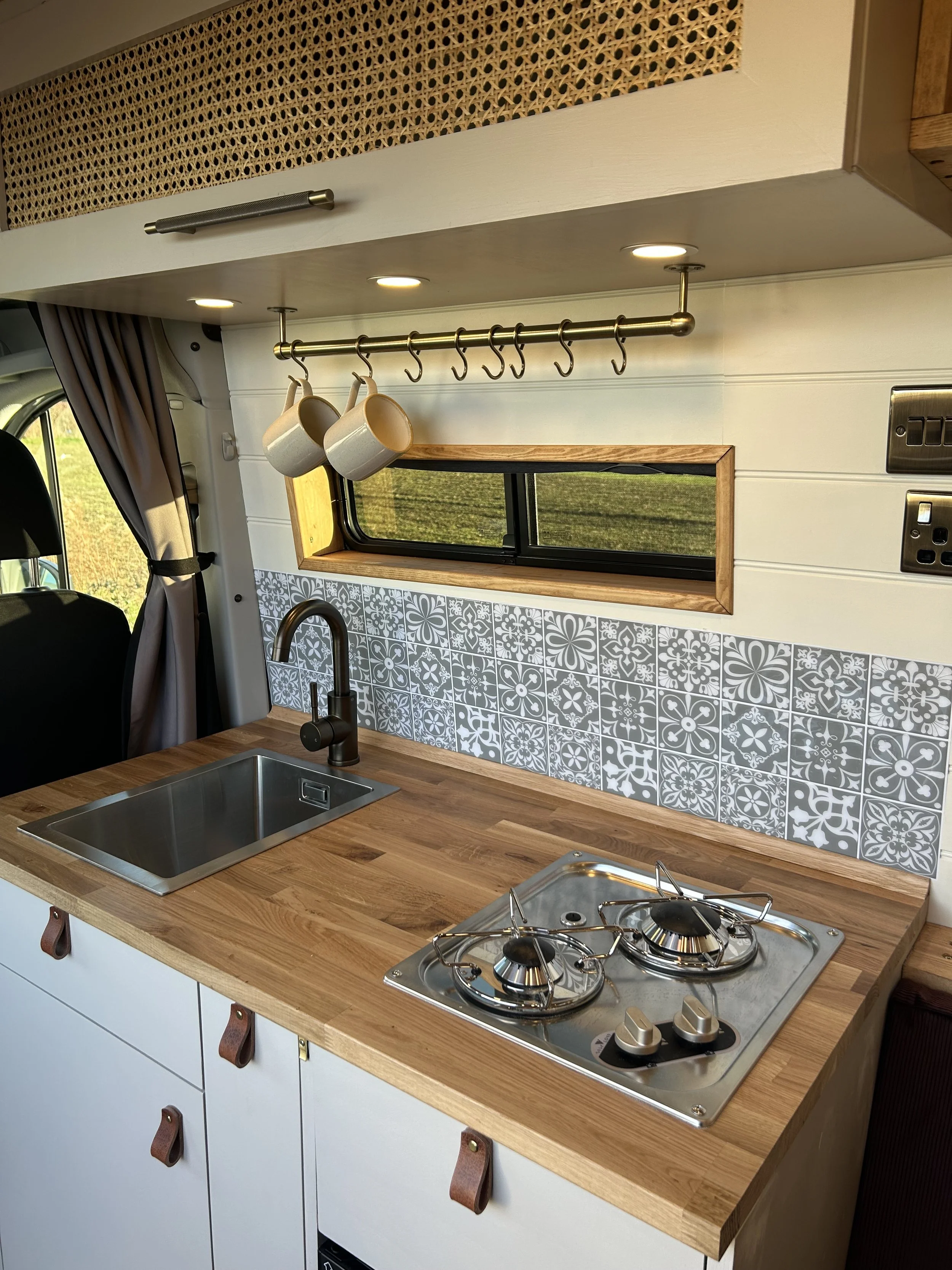 Compact kitchen area inside a camper van with a wooden countertop, a stainless steel sink, a two-burner gas stove, patterned wall tiles, a small window, hanging mugs, cups, and hooks, and some cabinetry.
