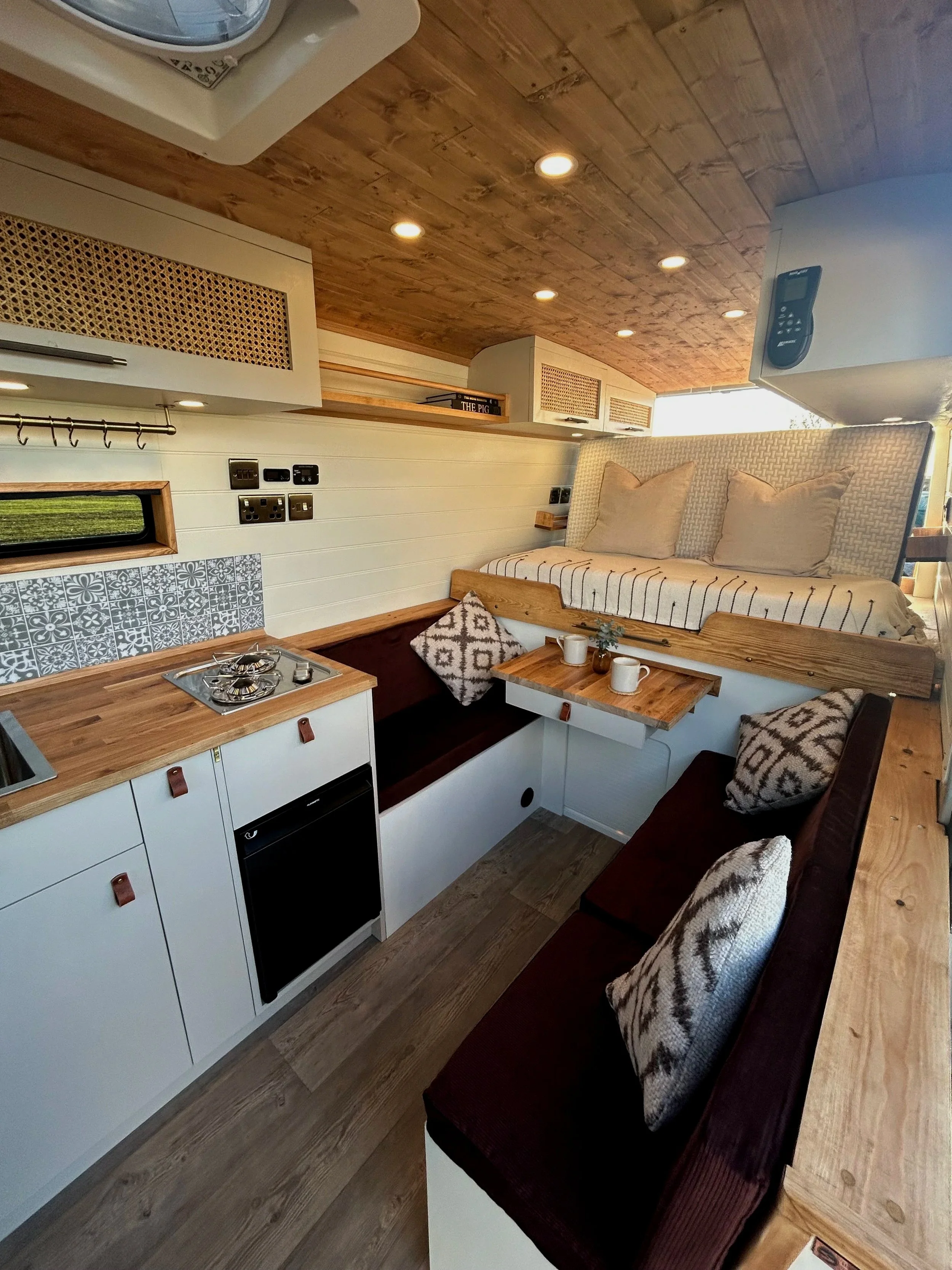 Interior of a compact camper van or tiny house with a wooden ceiling, white walls, a small kitchenette with a stove, and cozy seating area with pillows, a small table, and a sleeping loft with pillows.