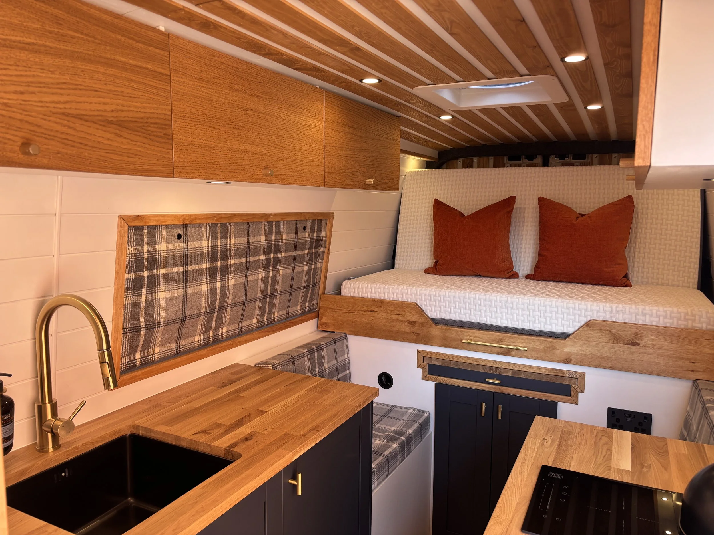 Compact living space with a cozy bed enclosed in a wooden frame, a cushioned bench with plaid fabric, a small kitchen area with a wooden countertop, a black sink, and a brass faucet. The ceiling has wooden beams and recessed lighting.