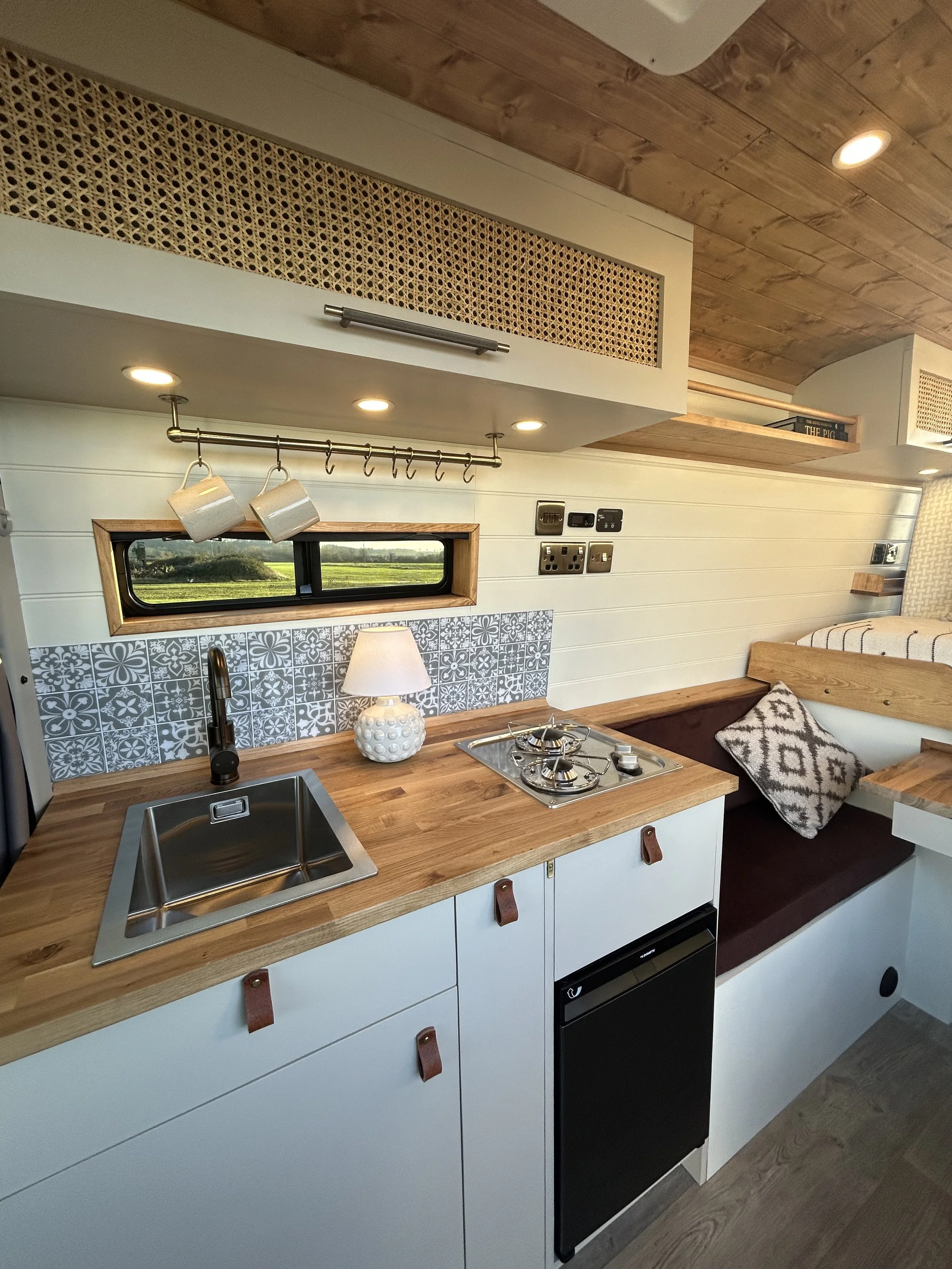 Interior of a compact kitchen and dining area with white cabinets, a wooden countertop, small sink, stove, and a window with a scenic view. There is a seating nook with a cushion and decorative pillow, and a small lamp on the countertop.