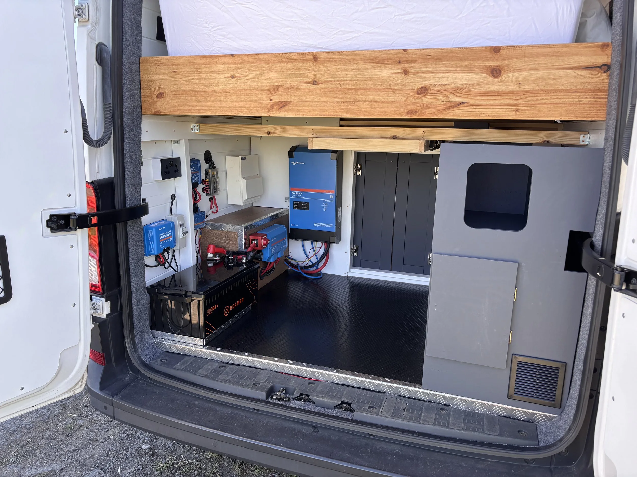 Inside a vehicle showing electrical setup with batteries, inverters, wiring, and wooden panels for organization.