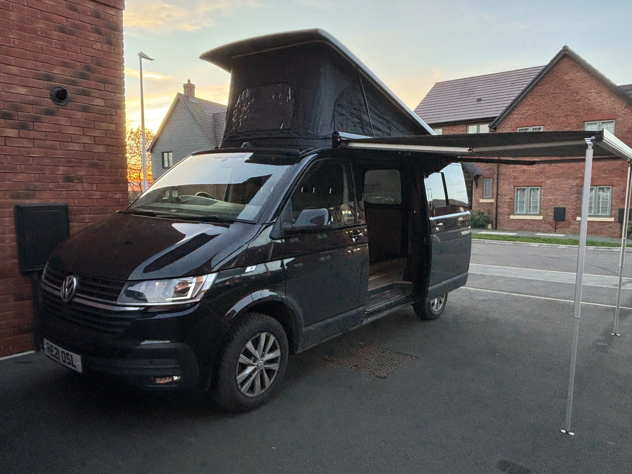 A black Volkswagen van with a pop-up roof tent and an extended canopy on a driveway in front of a brick wall and residential houses at sunset.