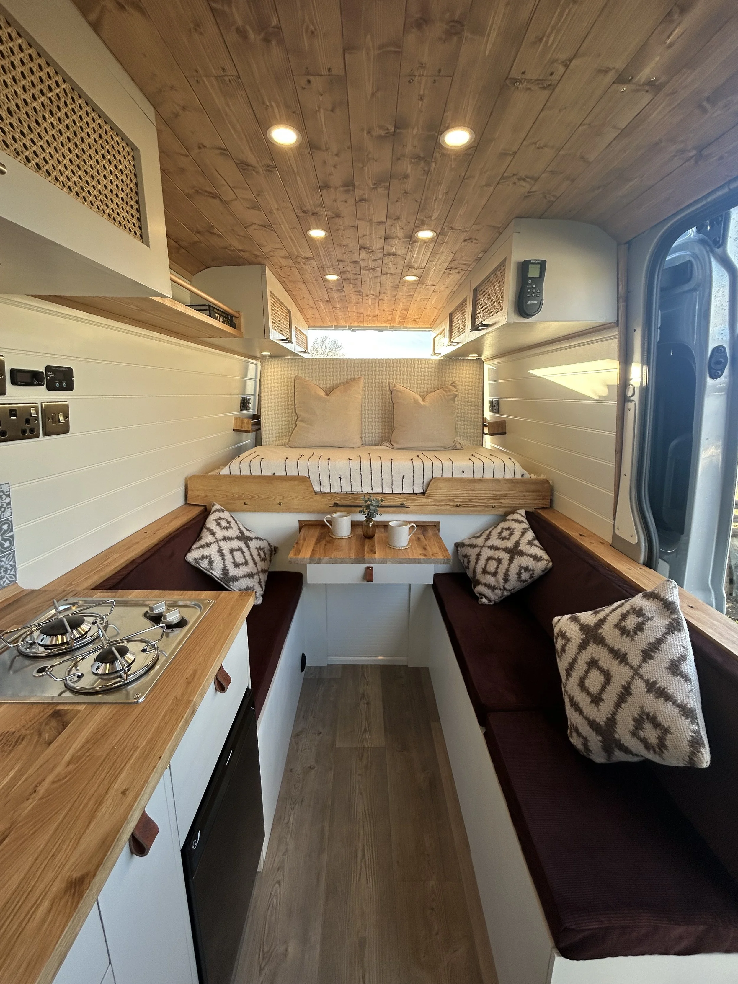 Interior of a tiny home on wheels with a kitchen area on the left, a seating nook with cushions on the right, and a sleeping area with pillows at the back, featuring wood paneling on the ceiling and walls.