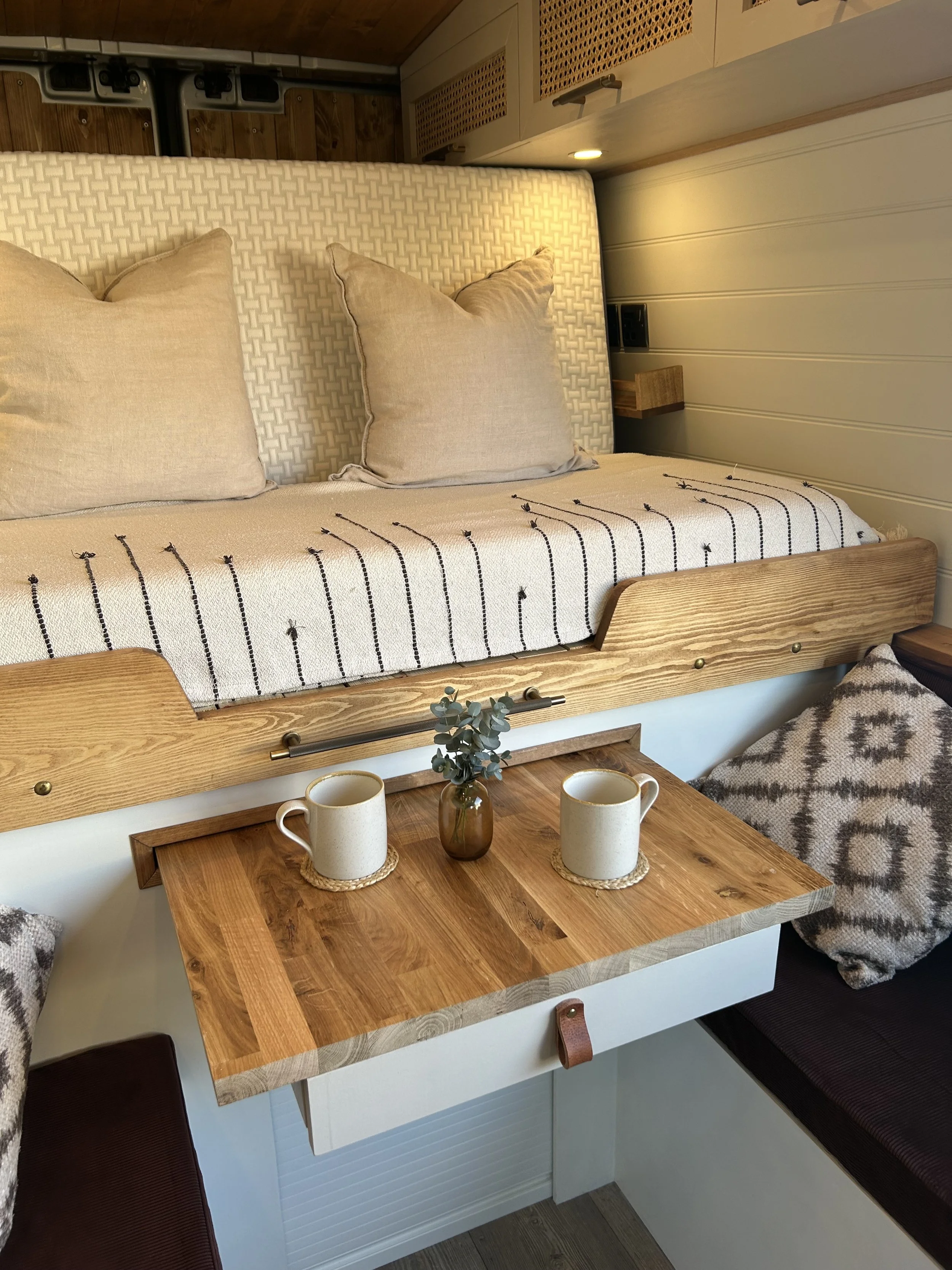 A cozy sitting area with a wooden table, two coffee cups on woven coasters, a small flower vase, and cushions; a bed with beige pillows and patterned blanket, with wood-paneled walls and small wall shelves in the background.