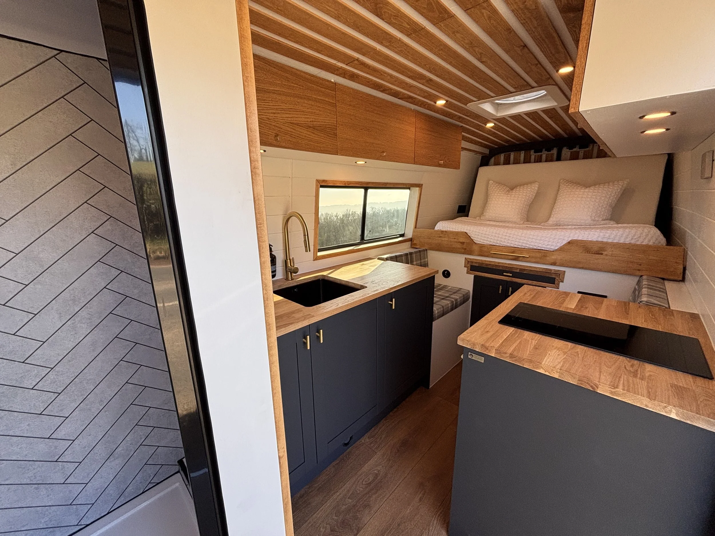 Small, modern kitchen and sleeping area in a tiny home with a bed above the kitchen cabinets, a window with a view, blue cabinets with gold handles, a wooden countertop, and a black sink.