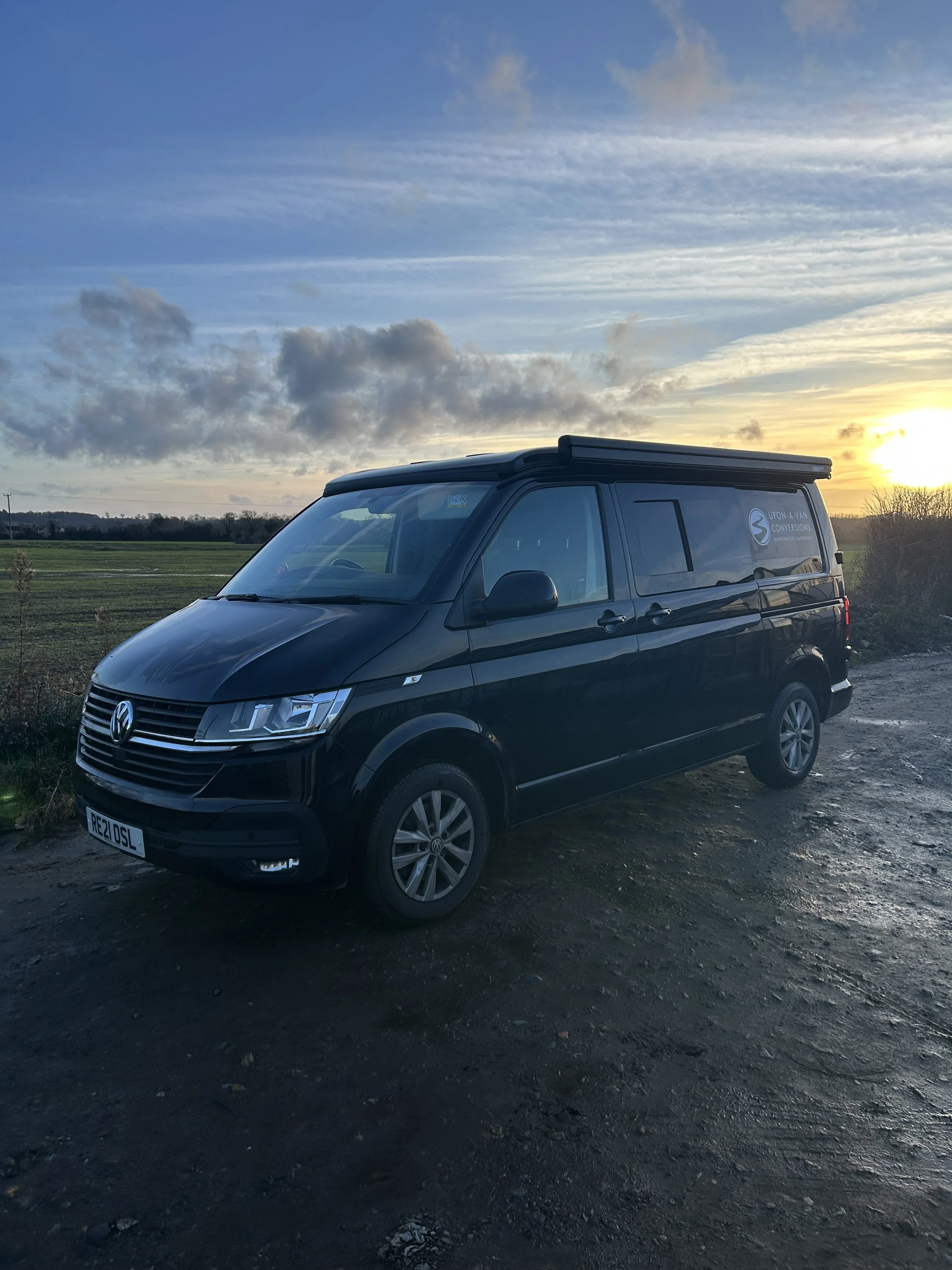 Black Volkswagen van parked on a gravel surface with a field and sunset in the background.