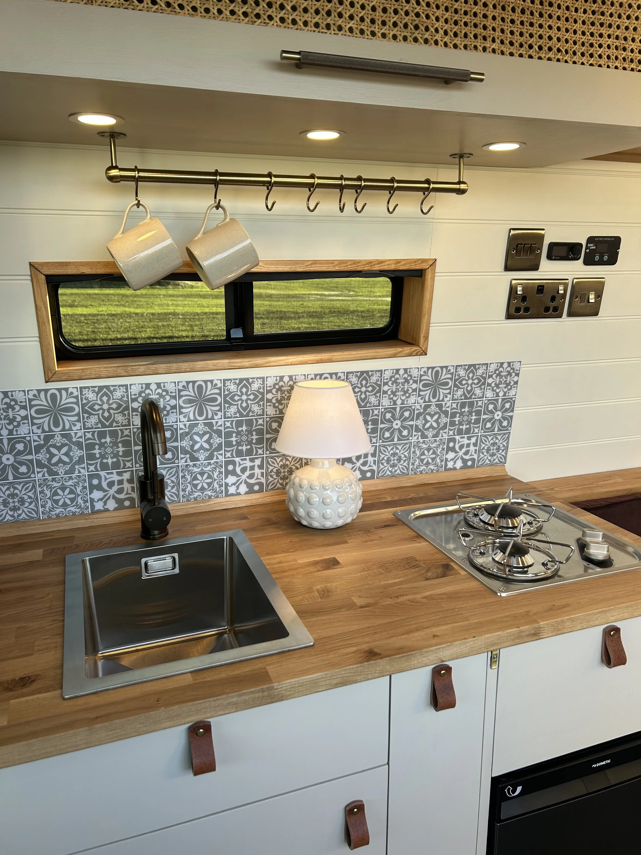 A compact kitchen with a wooden countertop, stainless steel sink, and a two-burner gas stove. Above the sink, there is a small window with a wooden frame, and a decorative patterned tile backsplash. Hanging above the countertop, there is a metal bar with hooks holding ceramic mugs. On the counter, there is a white ceramic lamp with a textured base.