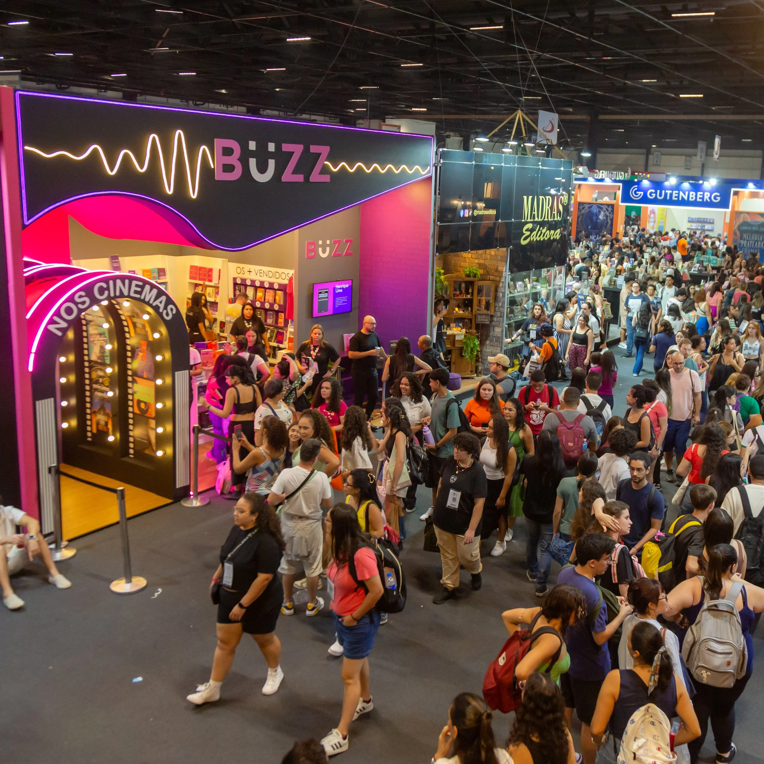 Imagem de uma feira com muitas pessoas caminhando entre estandes de livrarias e editoras, incluindo o estande da Buzz, com iluminação rosa e decorado com tema de cinema.