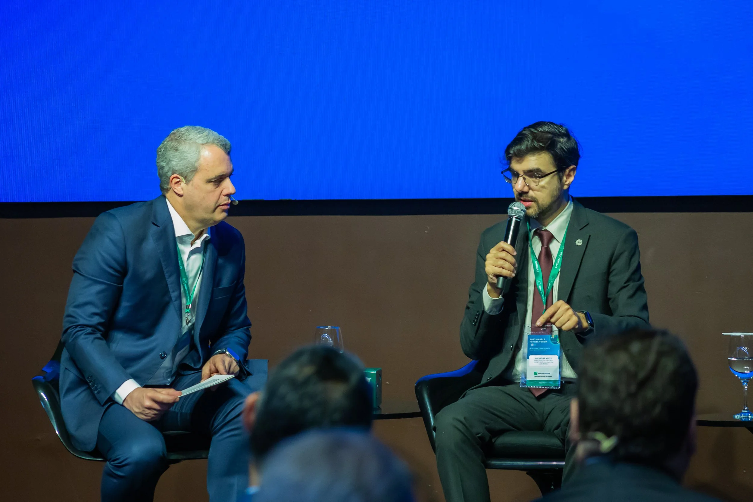 Duas pessoas em um painel de discussão, uma segurando um microfone, em um evento formal. Atrás delas, uma tela azul e público assistindo.