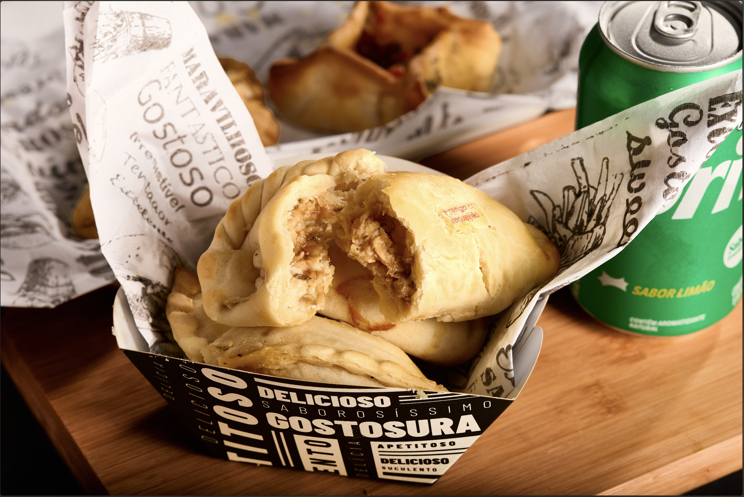 Empada de frango cortada ao meio em uma bandeja com papel decorativo que tem palavras relacionadas a gostosura, como 'delicioso', 'apetitoso' e 'sabor limão', ao lado de uma lata de refrigerante verde na madeira.