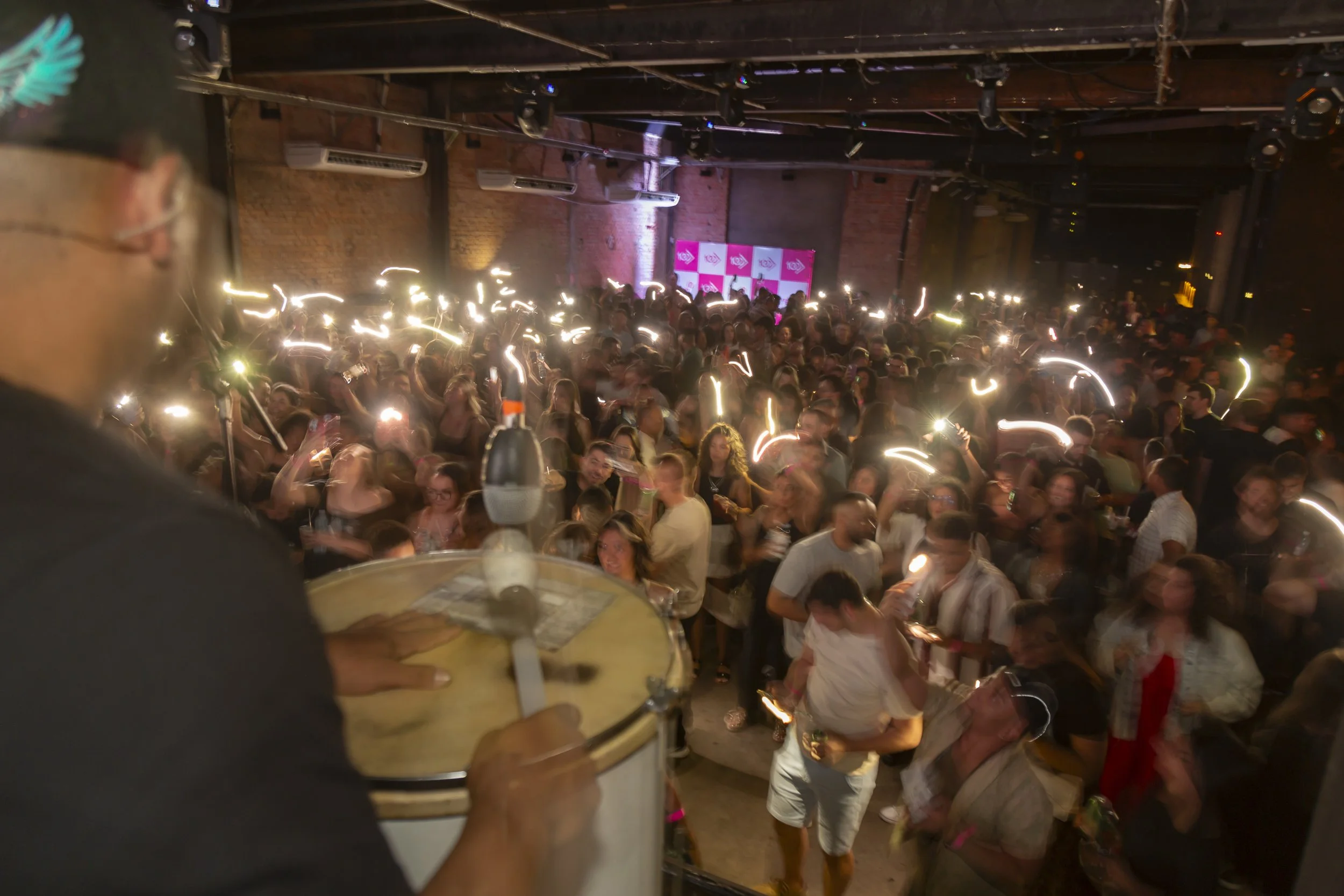 Pessoas em um evento noturno, com luzes brilhando enquanto seguram celulares. Em primeiro plano, uma pessoa tocando um tambor, com uma parede de tijolos ao fundo.
