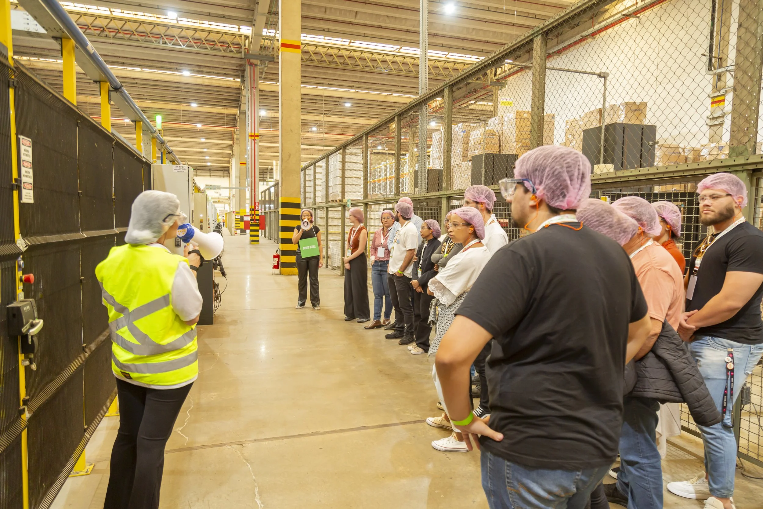 Grupo de pessoas recebendo instruções em uma fábrica, usando vestimentas de proteção como toucas, óculos e máscaras.