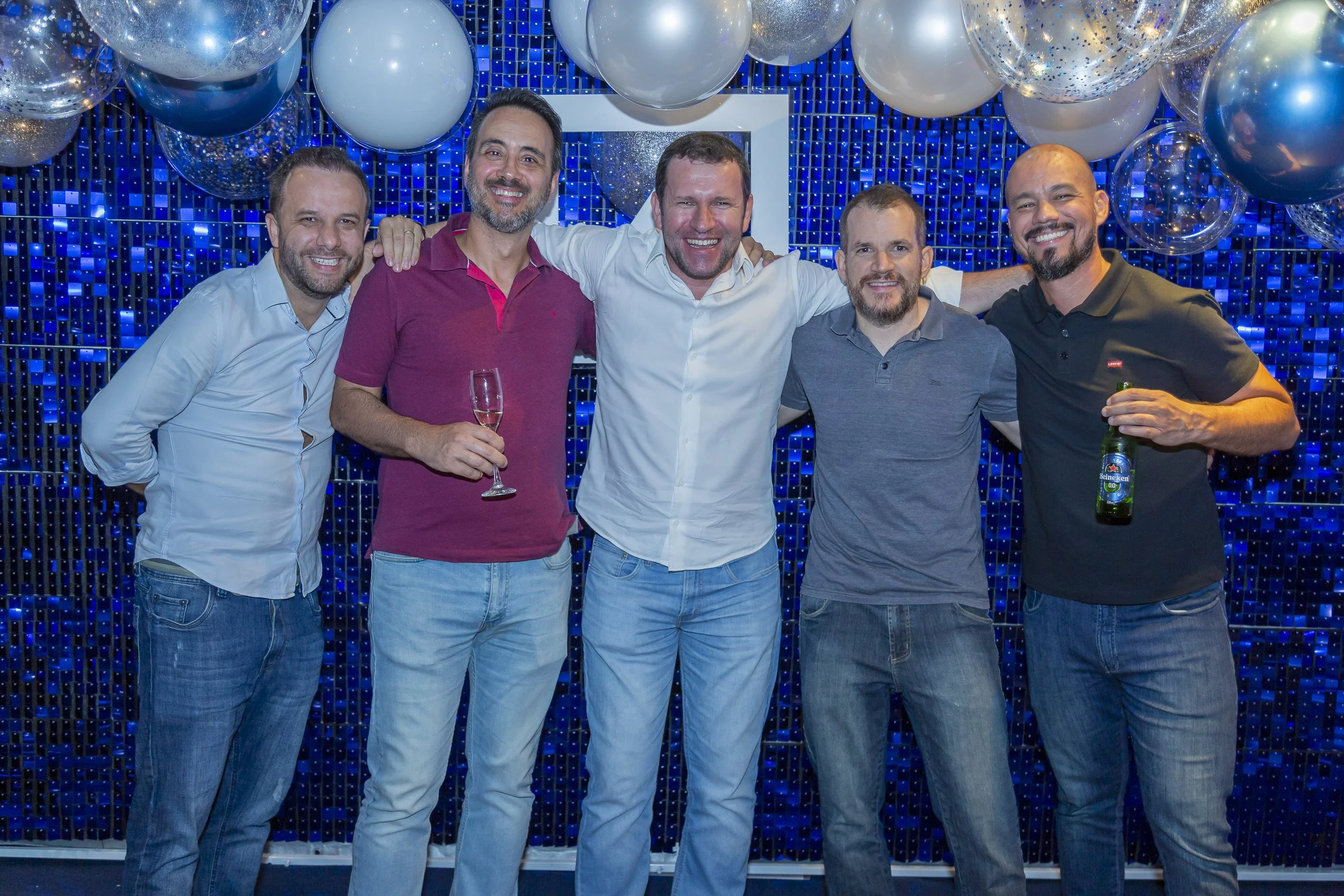 Grupo de cinco homens sorrindo, alguns segurando bebidas, em uma festa decorada com balões prateados e fundo azul de azulejos. 