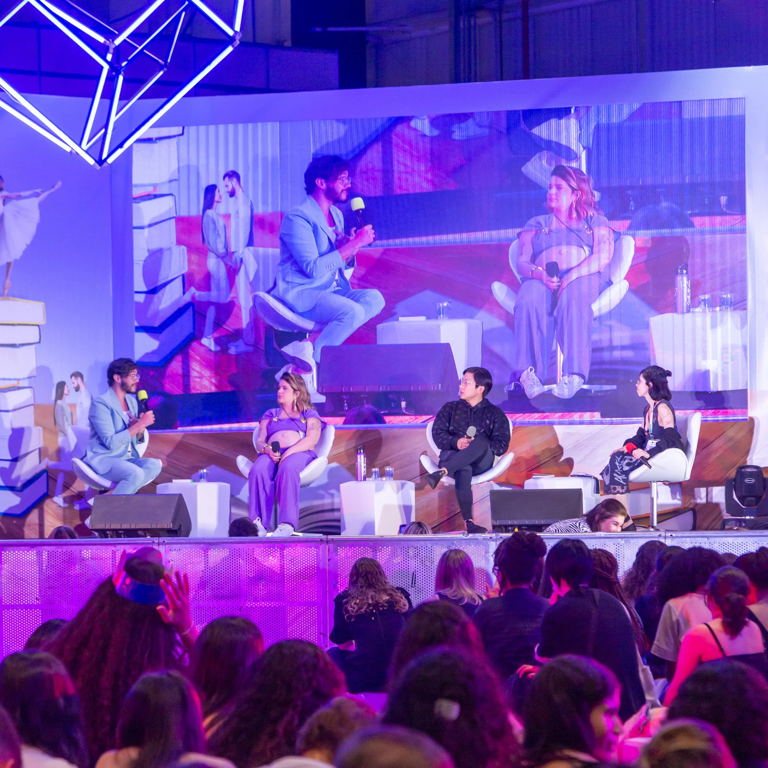 Painel de fundo de evento com quatro pessoas em uma mesa de debates, duas delas falando ao microfone e duas ouvindo, com uma plateia à frente. A cena é vista ao vivo e em uma projeção gigante atrás, com iluminação azul e roxa.
