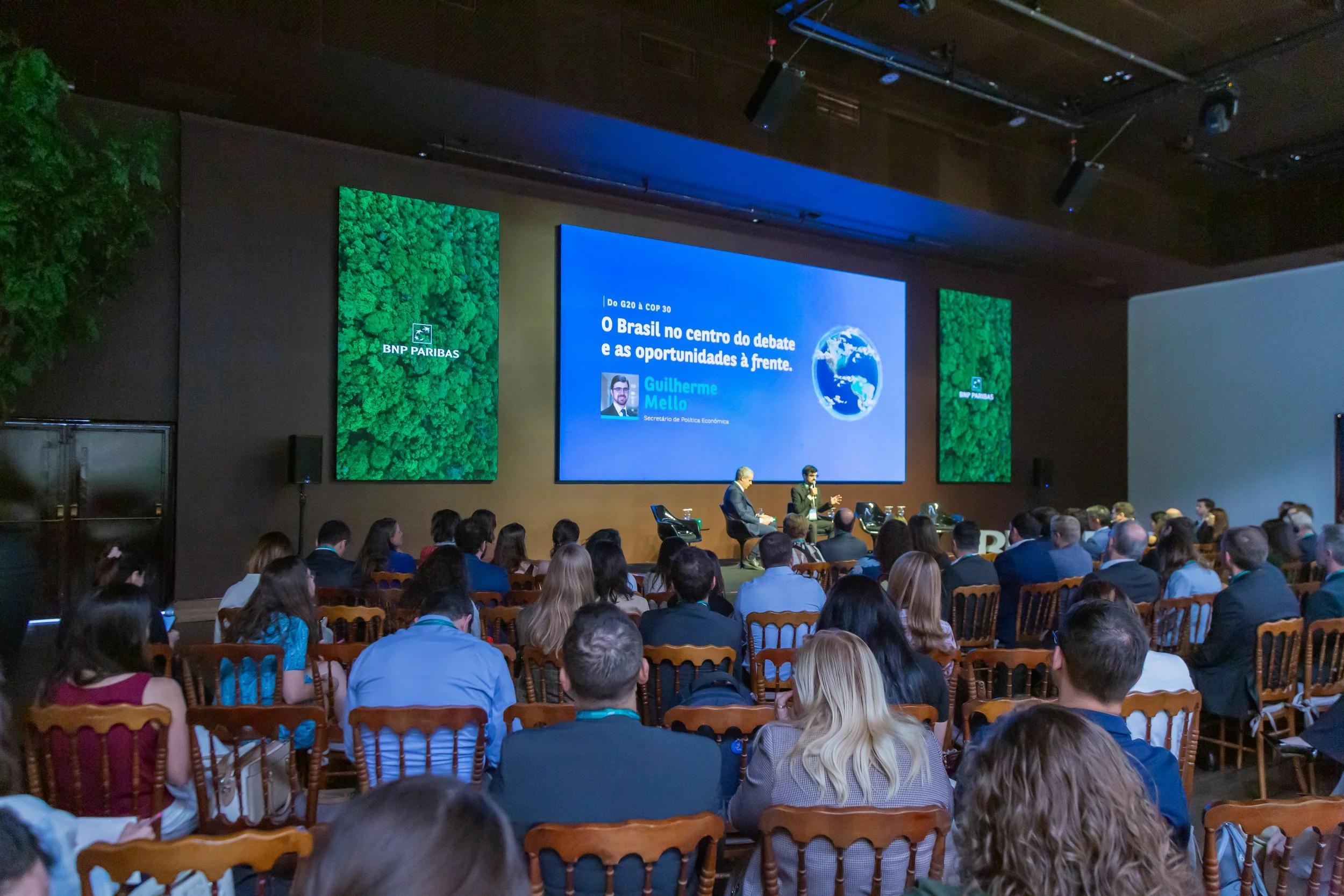 Palestra em um auditório grande com pessoas sentadas, uma tela grande ao fundo exibe o título "O Brasil no centro do debate e as oportunidades à frente" com a foto de Guilherme Mello.