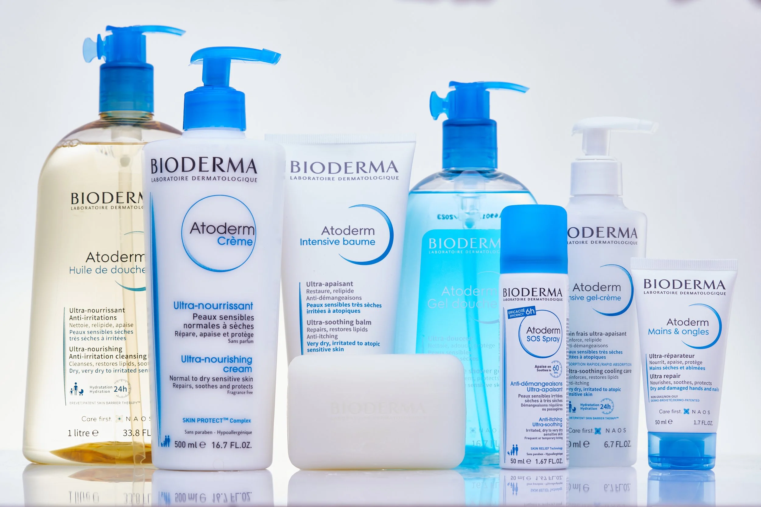 Diversos produtos de cuidados com a pele da marca Bioderma, incluindo sabonete líquido, creme, balm, gel de banho, spray anti-demangoes, creme para mãos e unhas em frascos de diferentes tamanhos, todos com embalagem branca e detalhes azuis.