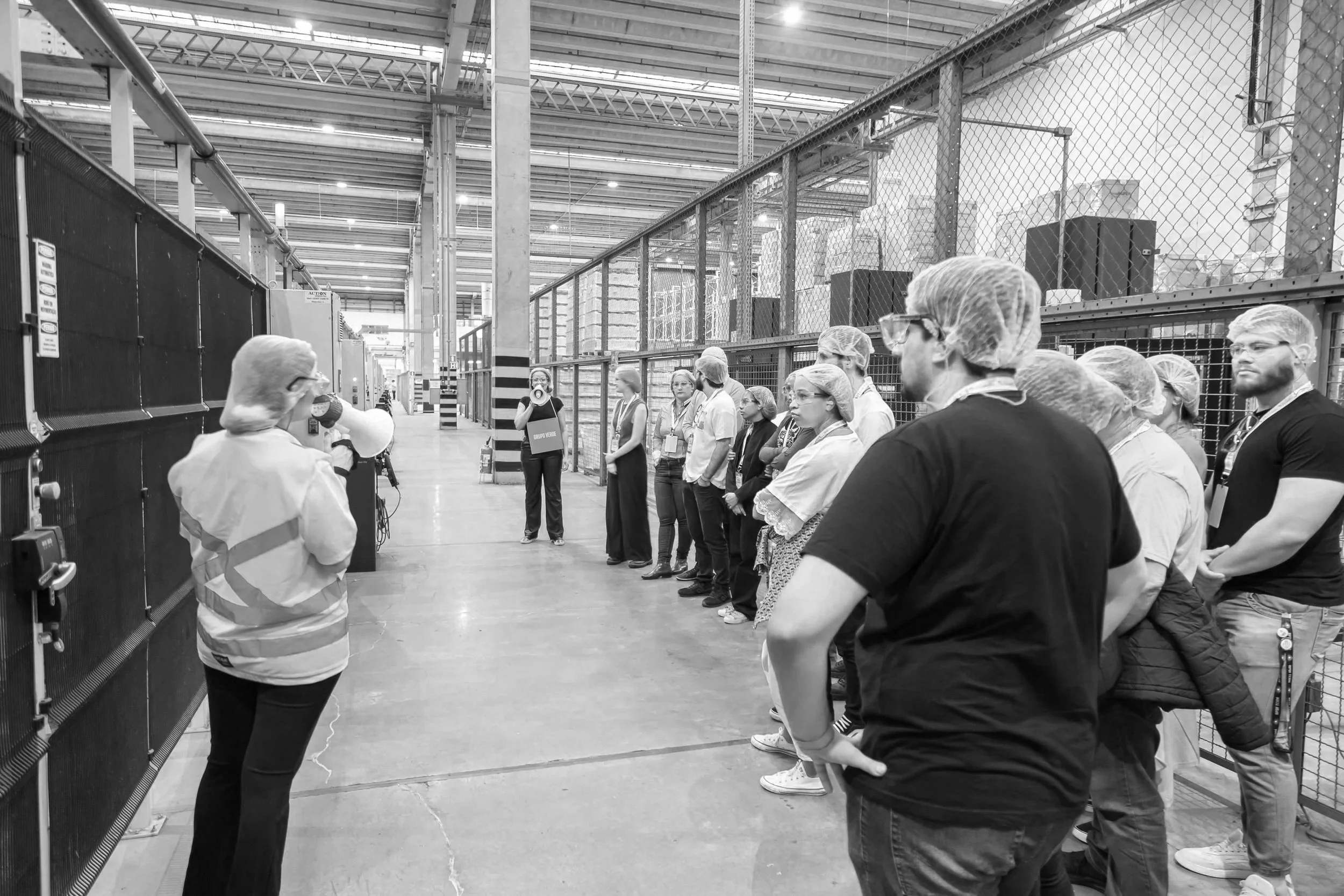 Grupo de pessoas em uma visita guiada dentro de uma fábrica, com cobrindo cabelo, ouvindo uma explicação de uma pessoa com megafone.
