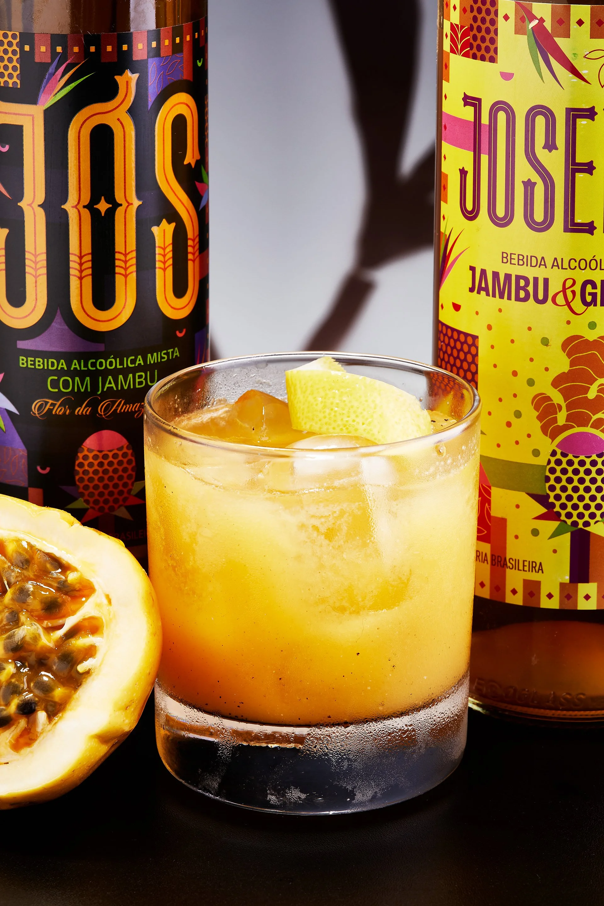 Copo com bebida alcoólica amarela com gelo, decorado com um pedaço de limão, ao lado uma metade de maracujá e duas embalagens de bebidas de sabor limão e maracujá.