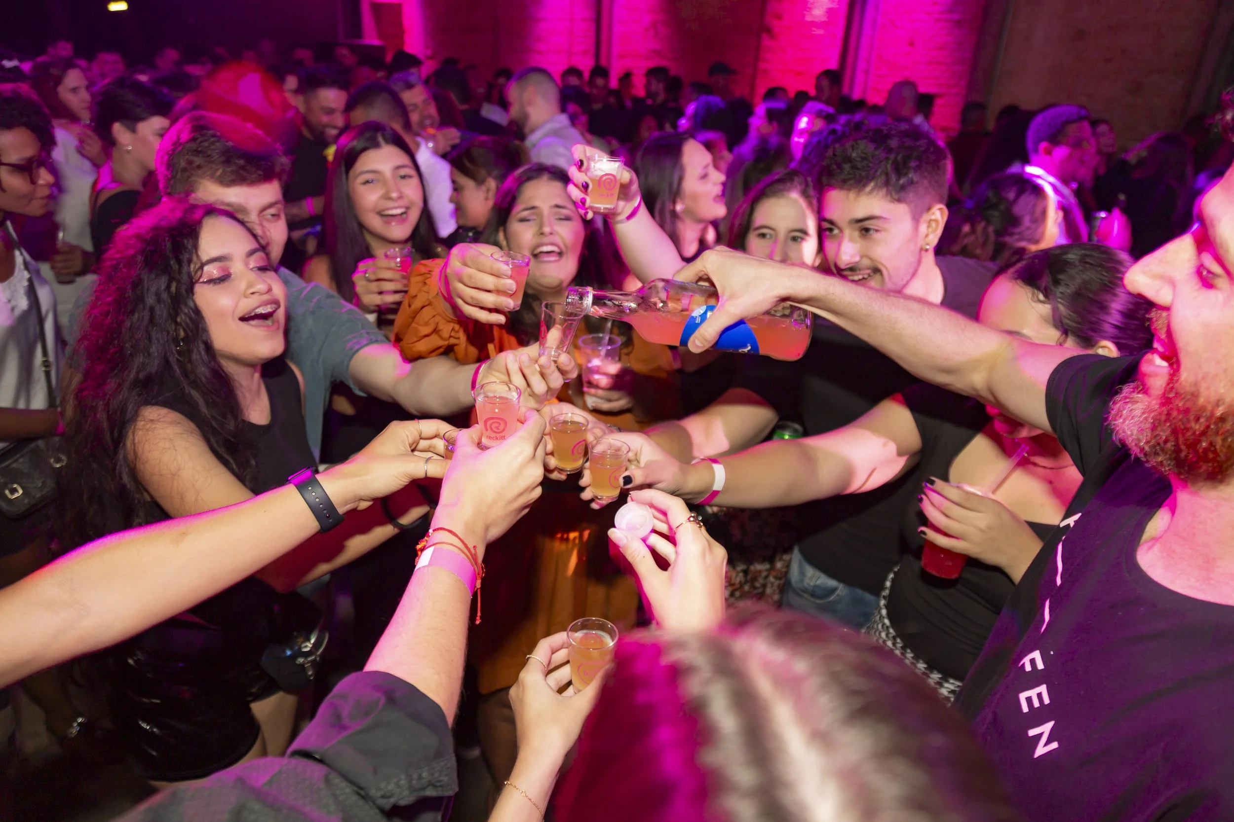 Grupo de pessoas celebrando com bebidas em um ambiente com luz neon rosa, muitas pessoas sorrindo e brindando juntas.