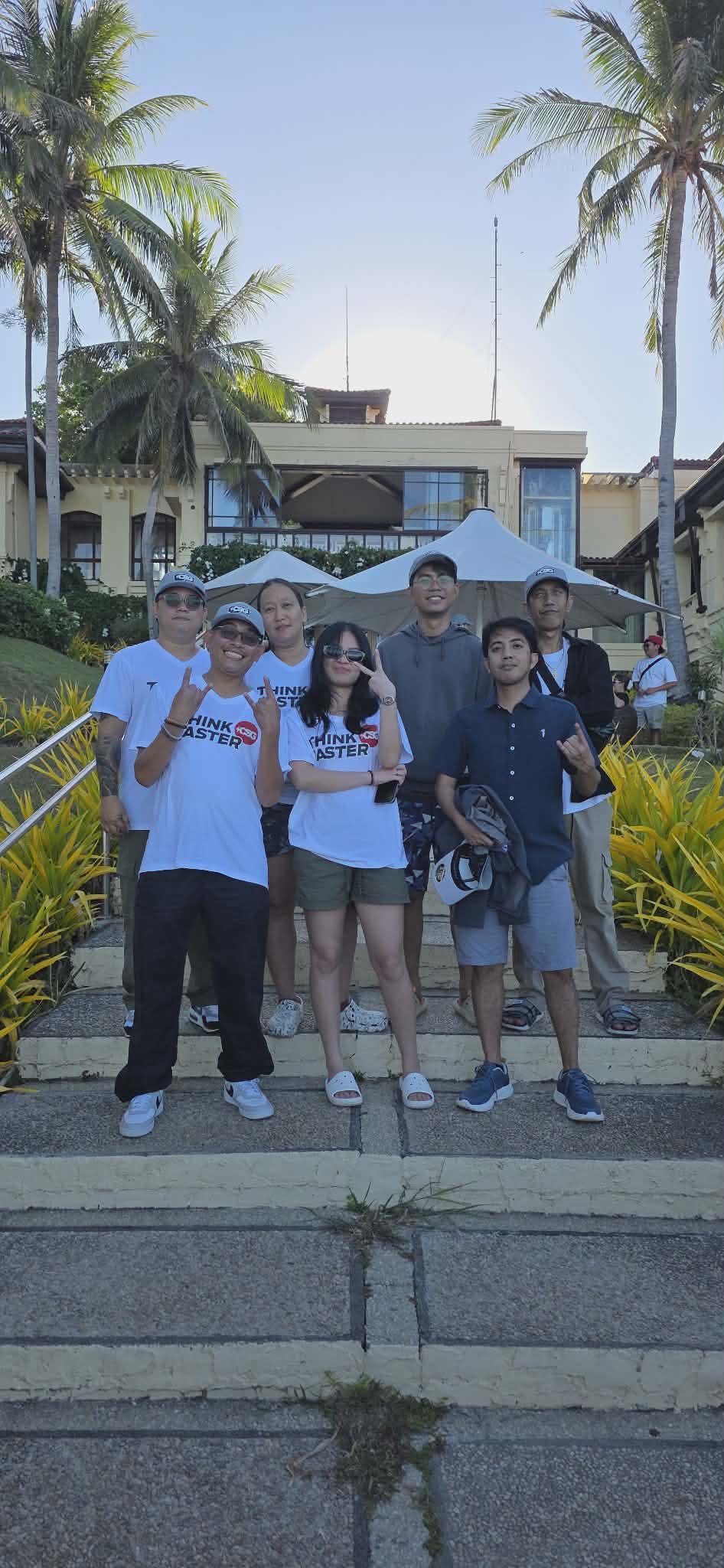 Group of seven people standing on steps outside a building with palm trees and tropical plants, some making hand gestures, wearing casual summer clothing, with umbrellas and a hotel or resort in the background, and the sun setting behind the building.
